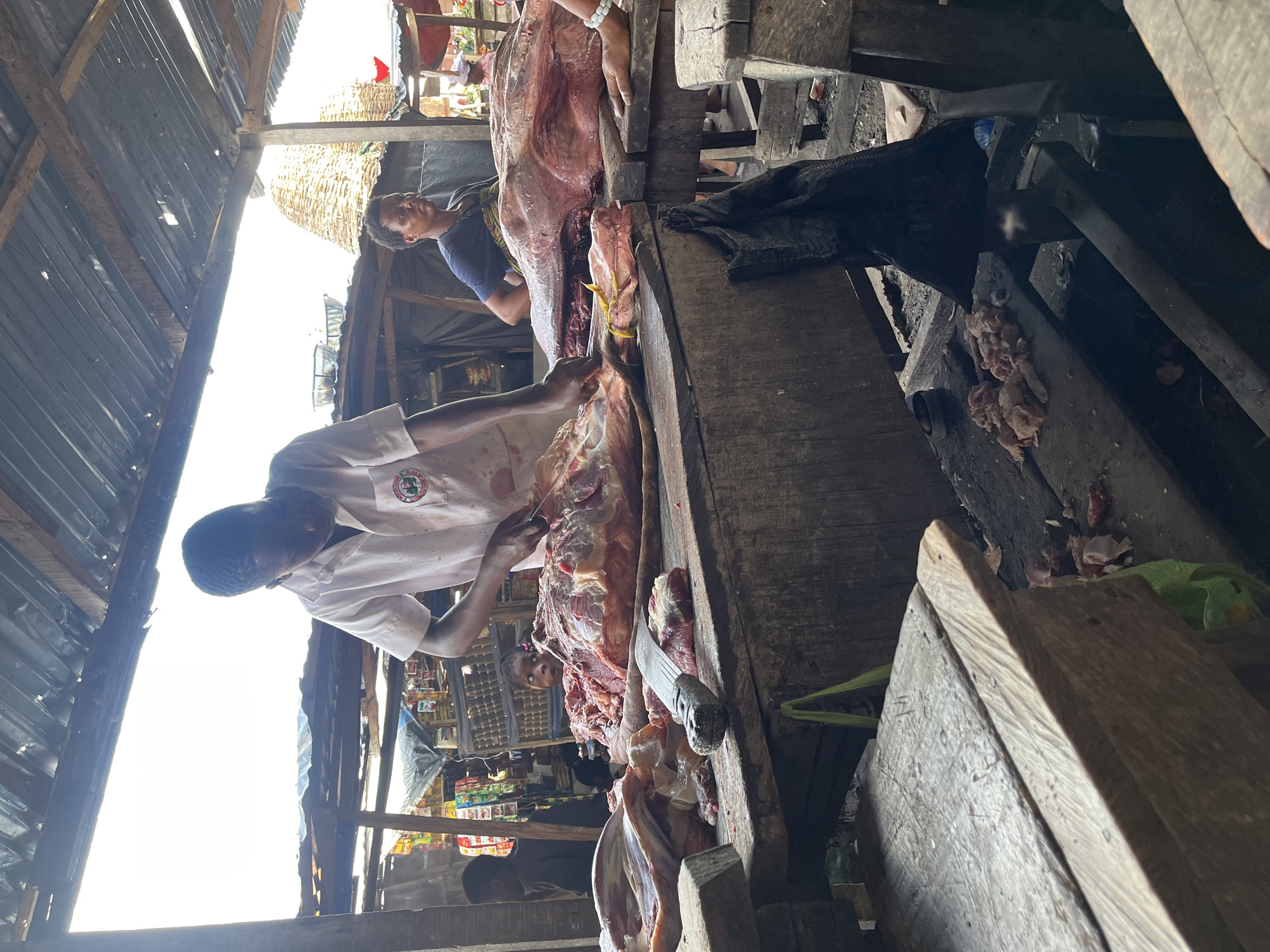 Ado, Nigeria: A man in a white shirt is butchering meat on a wooden table. Another man watches. The scene is in an open-air market under a corrugated metal roof with wooden beams. There are shops with goods and people in the background. A cleaver rests on the table. Cut pieces of meat are scattered about.