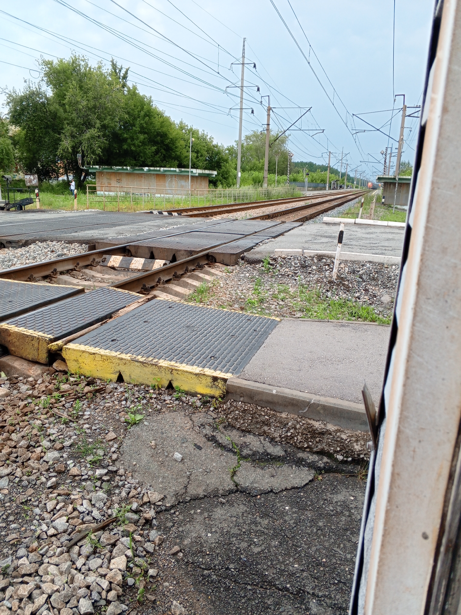 The image depicts a railroad crossing in Kamensk-Uralsky, Russia, on an overcast day. The foreground features several sets of train tracks with metal crossing plates, some of which are painted yellow. The ground around the tracks is covered with gravel and patches of grass. In the middle ground, a person is visible standing near a small building with a green roof. The background shows more train tracks, overhead electrical wires supported by poles, and dense green trees. Further in the distance, a train with an orange locomotive is approaching. The perspective is from a low angle, looking along the tracks.