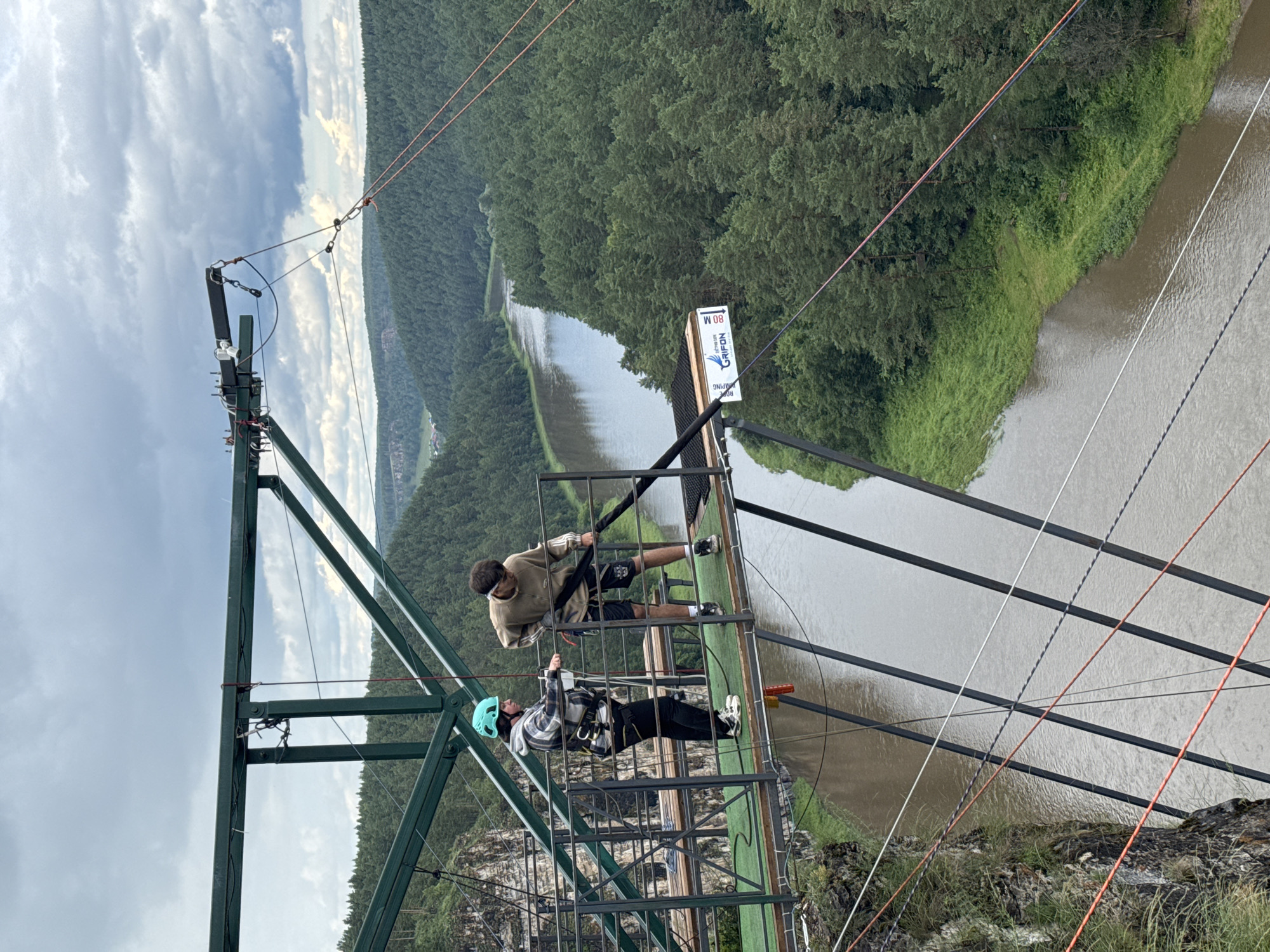 The image shows a bungee jumping platform on a metal structure overlooking a wide, tree-lined river in Kul'metovo, Russia. Two individuals are on the platform. One person, wearing a blue helmet and a checkered jacket with a safety harness, stands near the railing. The other person, wearing a light brown hoodie and dark shorts, is holding a thick black rope, appearing to prepare for a jump. The river below is flanked by dense green forest, and the sky is overcast with dramatic clouds. The platform is equipped with safety features like railings and ropes. A sign on the platform indicates "Grifon" and "80 M," suggesting it is a bungee jumping site with a significant drop.