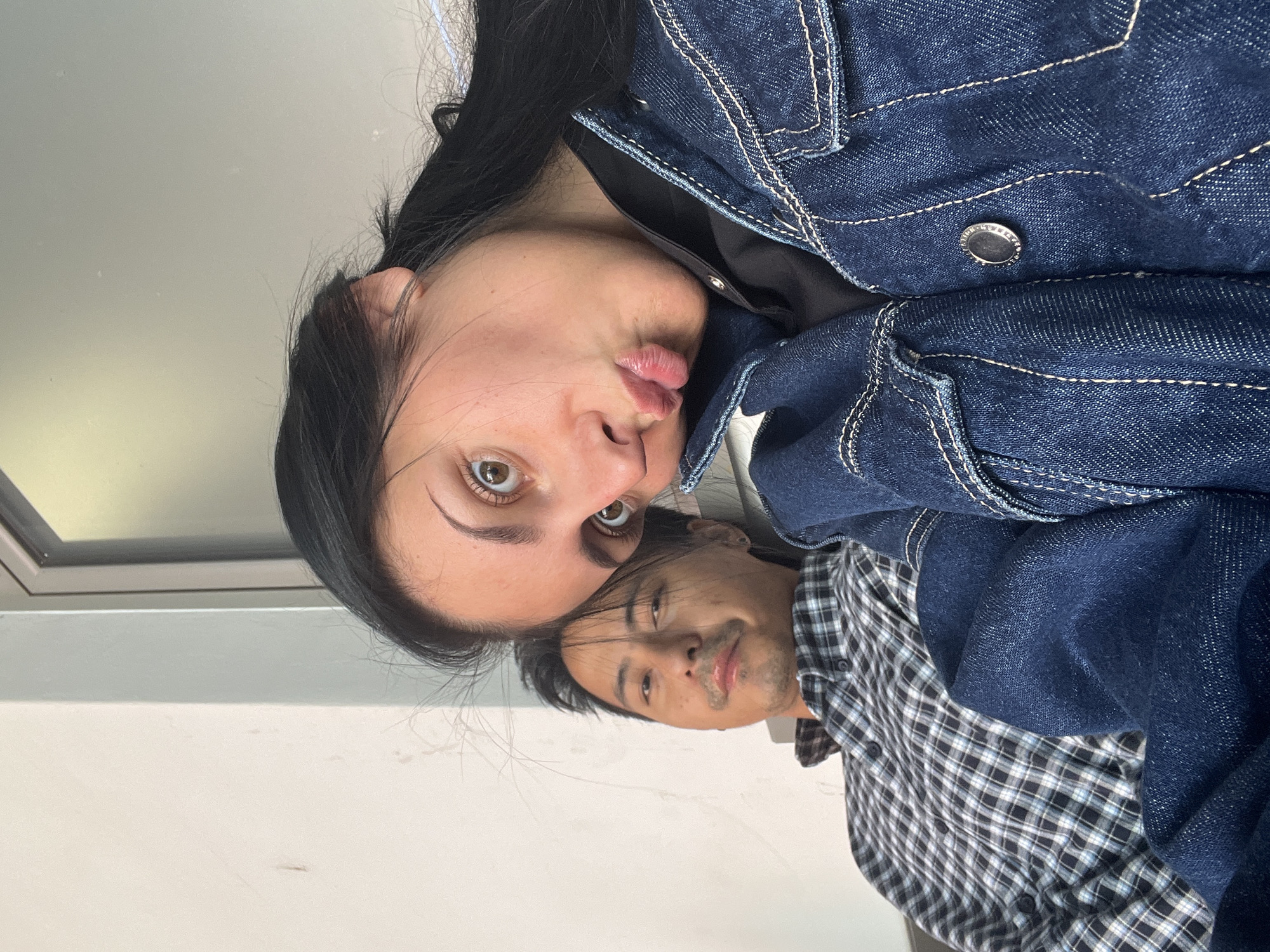 Two people, a woman and a man, are positioned closely together, presumably taking a selfie. The woman, in the foreground, is wearing a dark blue denim jacket over a black top and has dark hair pulled back. She is making a duck face expression. Behind her, the man is wearing a plaid shirt in black, white, and blue tones. The background is a blurry mix of white and grey, suggesting an indoor environment. The angle of the shot is slightly off-kilter. There is no visible information that identifies the exact location in Sofia, Bulgaria.