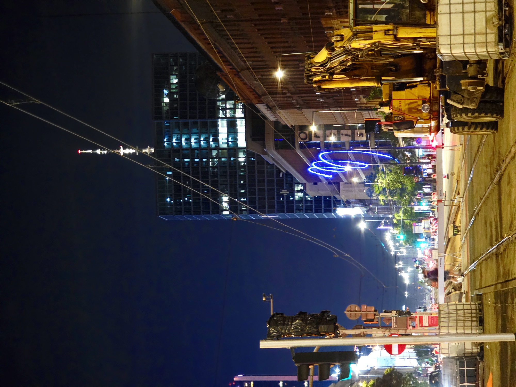 The image depicts a nighttime street scene in Warsaw, Poland, likely on or near Marszałkowska Street, a major thoroughfare known for its construction and ongoing urban development projects. A large, yellow excavator is parked in the foreground on the right side of the frame, its mechanical arm partially visible. The street is under construction, with tram tracks visible and barriers lining the road.
In the background, a tall, modern skyscraper is illuminated, its windows lit from within, suggesting activity within the building. The sky is dark blue, indicating nighttime. Several traffic lights are visible, some showing green, others red. The presence of construction equipment and materials suggests ongoing infrastructure work.
There are a few individuals visible in the scene, appearing to be pedestrians or construction workers, though their actions are not clearly defined. The overall atmosphere is one of a bustling urban environment undergoing significant change, captured at night with artificial lighting creating dramatic contrasts. The architecture suggests a mix of older, brick buildings and modern high-rises. Electrical wires and streetlights crisscross the sky, further emphasizing the urban setting.
