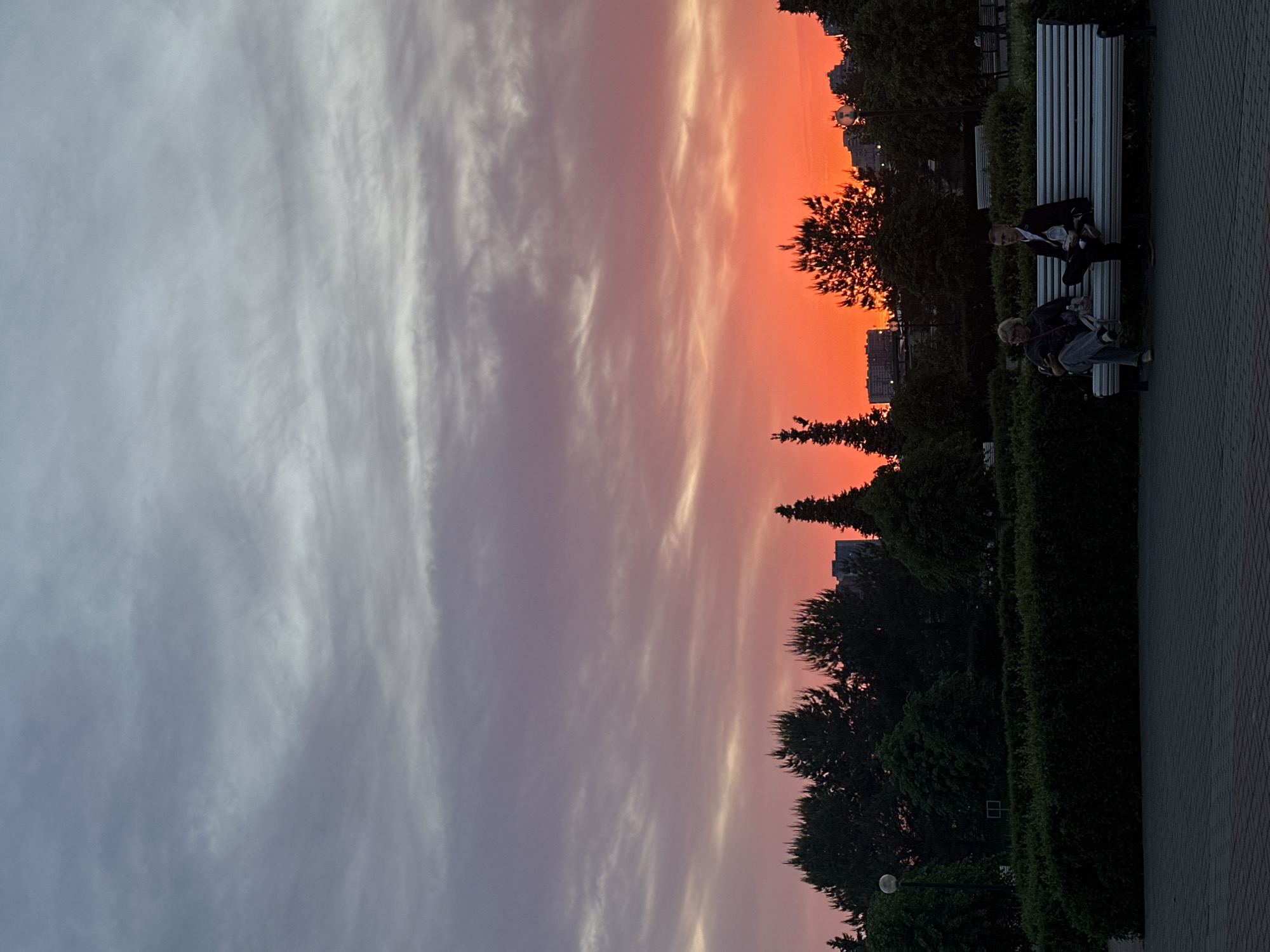 The image depicts a sunset in Kazan, Russia. The sky transitions from gray-blue on the left to orange on the right. Wispy clouds are visible across the sky. Trees and buildings are silhouetted against the colorful sky. On the right side of the image, two people are sitting on a white bench along a path. The path is made of patterned paving stones. A hedge separates the path from the trees. A lamppost is also visible near the bottom right corner.