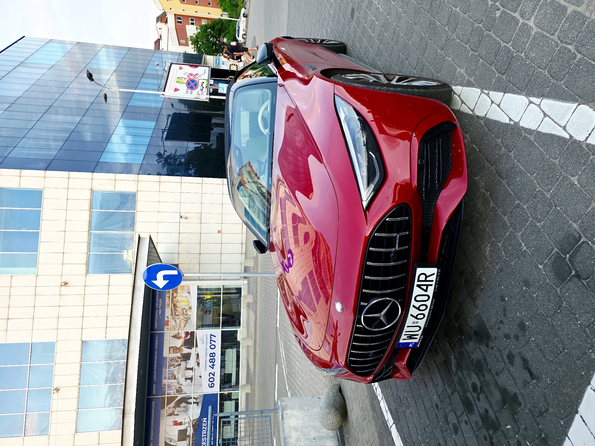 A red Mercedes-Benz convertible is parked in a lot with a modern glass-fronted building in the background. The car's front grille, headlights, and license plate are visible. The license plate reads "WU 6604R". A blue directional sign indicating a left turn is mounted on a pole to the left of the building. The ground is paved with cobblestones. In the background, a street lamp and other buildings are visible. Two people are walking in the distance on the right side of the image. The sky is overcast.