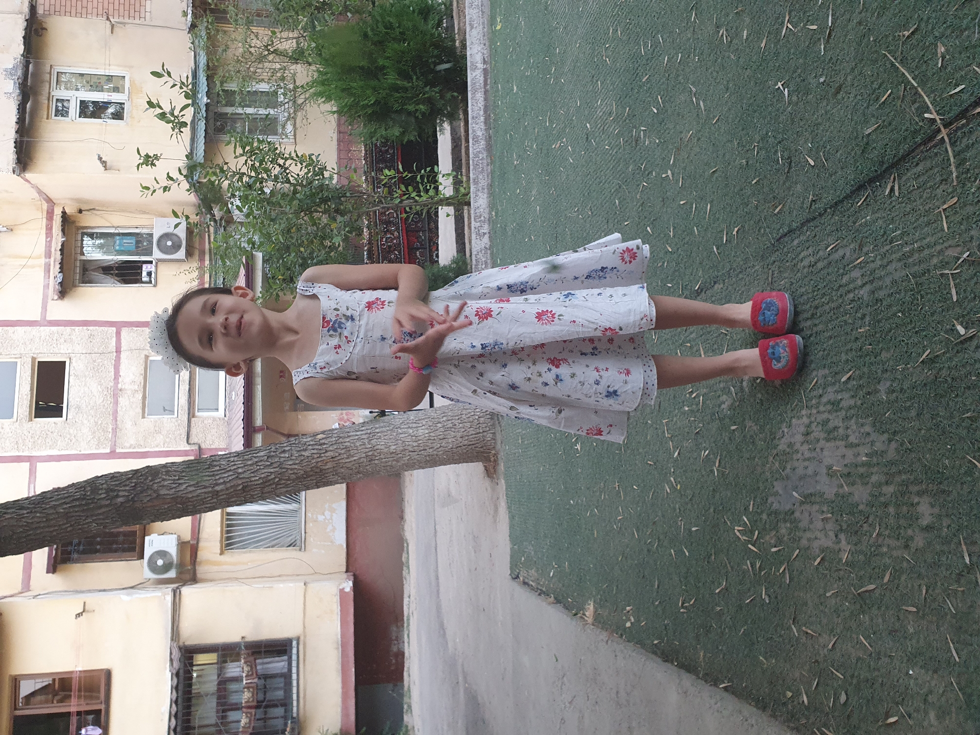 The image captures a young girl standing on a green, artificial turf surface with scattered brown leaves. She is wearing a white floral print dress with red and blue flowers and red slippers with a blue design. A tree trunk is prominent in the background, to the left of the girl, with its bark textured and rough. Behind the tree, a faded yellow building with brown trim and several windows is visible. Some windows have bars, and one has an air conditioning unit attached. The building's facade shows signs of wear and tear, with chipped paint. Further to the right, another building, lighter in color and with red brick accents, can be seen. Green bushes and trees are also present in the background, suggesting an outdoor urban setting. The overall scene is consistent with an apartment complex courtyard in Tashkent, Uzbekistan, given the architectural style and presence of common urban flora.