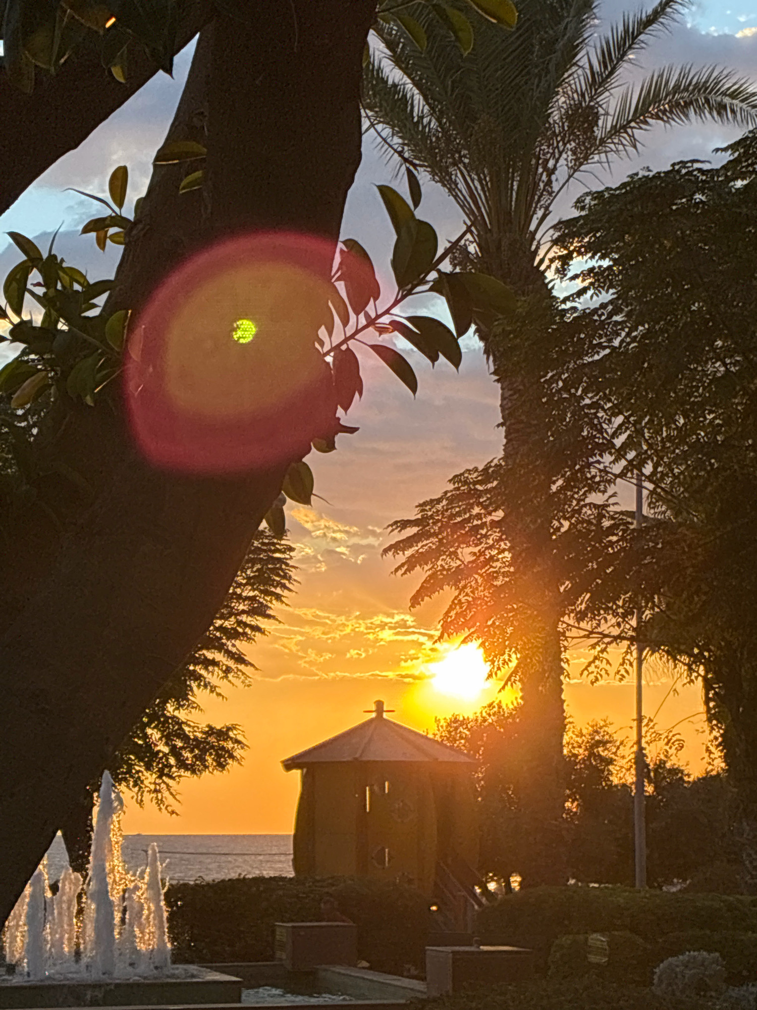 A sunset scene in Alanya, Turkey, features a fountain in the foreground with water jets. A dark tree trunk is to the left, with leaves and branches visible. A distinct lens flare with a circular halo surrounds a bright green circle, likely from the sun. In the midground, a small, dark, octagonal gazebo with a cross-shaped finial stands near a body of water. Palm trees and other foliage frame the view. The sun is setting over the sea, casting an orange glow across the sky and clouds.
