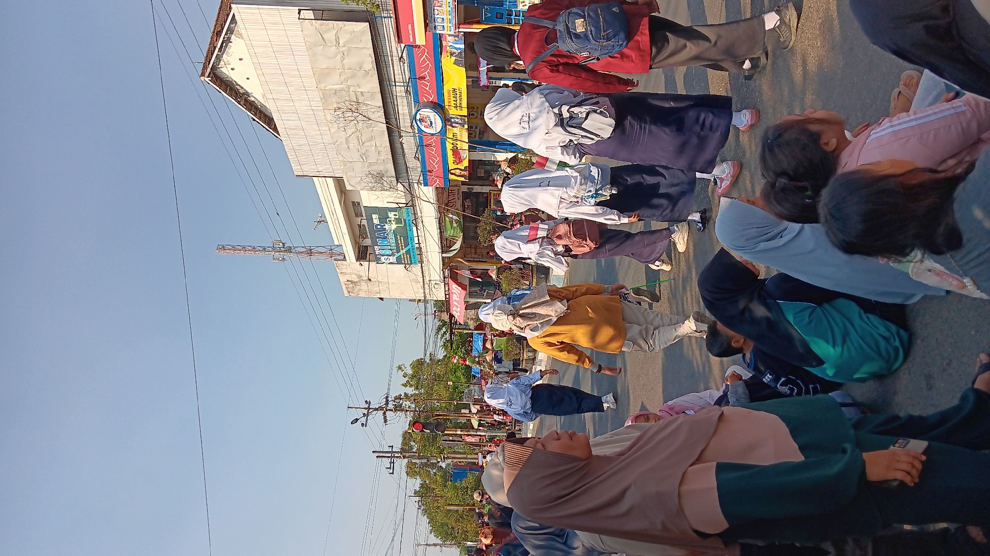 In Warujayeng, Indonesia, a street scene unfolds under a clear blue sky. A group of people, predominantly young women and girls wearing hijabs and school uniforms, are gathered on a paved road lined with buildings. Some are walking, while others are seated or standing. The buildings exhibit typical urban architecture, with shops and signage, including one that appears to be for an electronics store. A traffic light, currently red, is visible, indicating a regulated intersection. Overhead, power lines and a telecommunications tower are prominent features of the urban landscape. The overall atmosphere suggests a public gathering or event taking place on a sunny day in this Indonesian city.