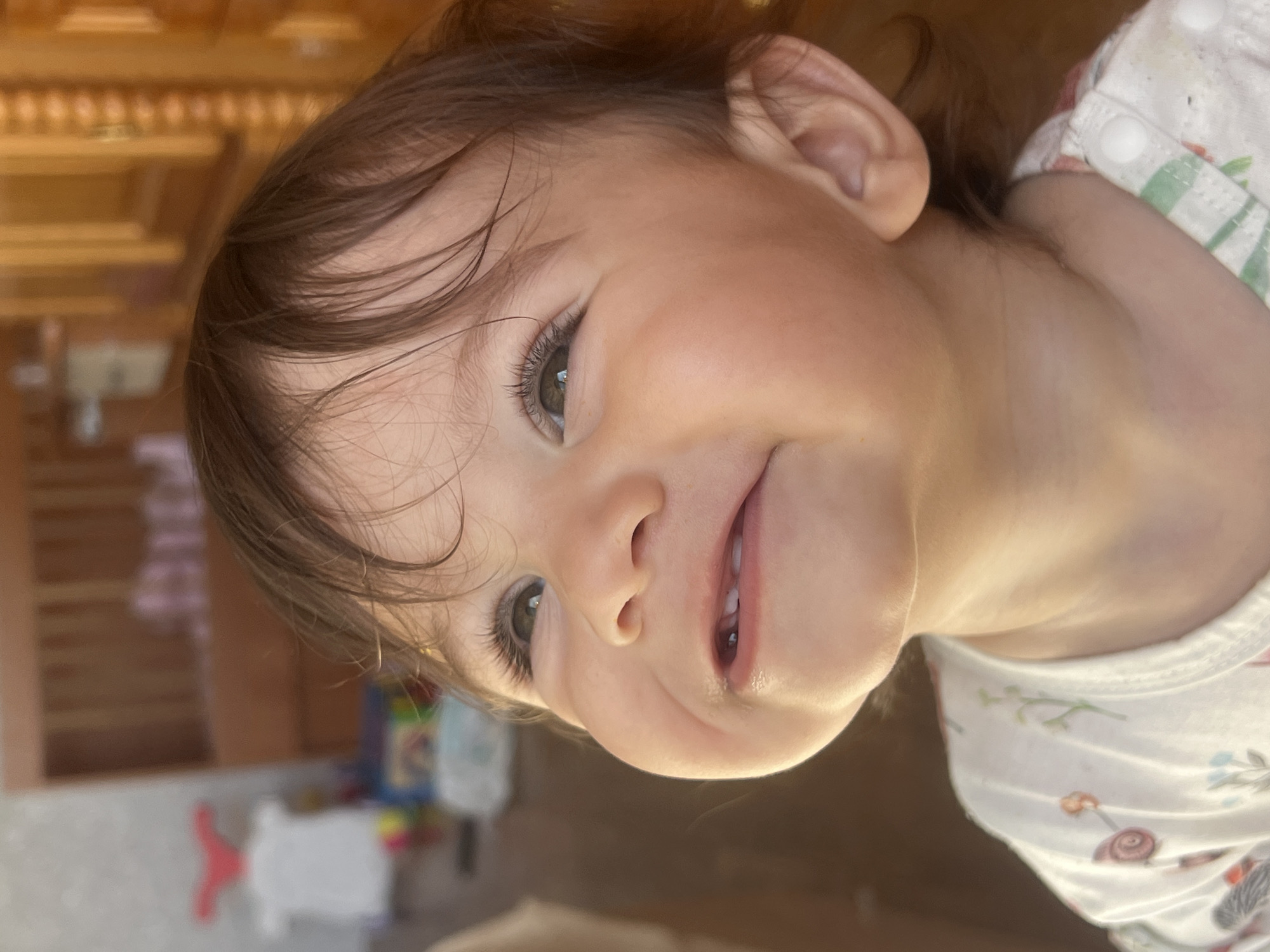 This close-up portrait captures a young child with a joyful expression, tilting their head. The child has shoulder-length, dark brown hair with strands falling across the forehead. Their bright, light-colored eyes (likely green or hazel) are wide open, looking directly towards the viewer. A gentle, open-mouthed smile reveals two small bottom teeth, with a hint of moisture on the lower lip. The child's skin appears smooth and healthy, well-lit with natural, soft light that highlights the cheekbones.
The child is wearing a white garment, possibly a top or a onesie, adorned with a delicate, colorful pattern that includes floral motifs and small, stylized animals like snails and mushrooms. Visible parts of the left ear are also seen.
The background is significantly blurred, indicating a shallow depth of field, which keeps the focus entirely on the child. The indistinct background features warm, earthy tones, suggesting an indoor setting with possibly wooden furniture or walls. Some out-of-focus shapes and colors are discernible, such as a bright red and white object and a stack of colorful items, hinting at toys or household objects.
The overall emotion conveyed is happiness and contentment. The bright, even lighting suggests it is daytime. While the specific location cues are not visible in the image itself, the provided context indicates the photograph was taken in Elena, Bulgaria. There is no visible text in the image.
