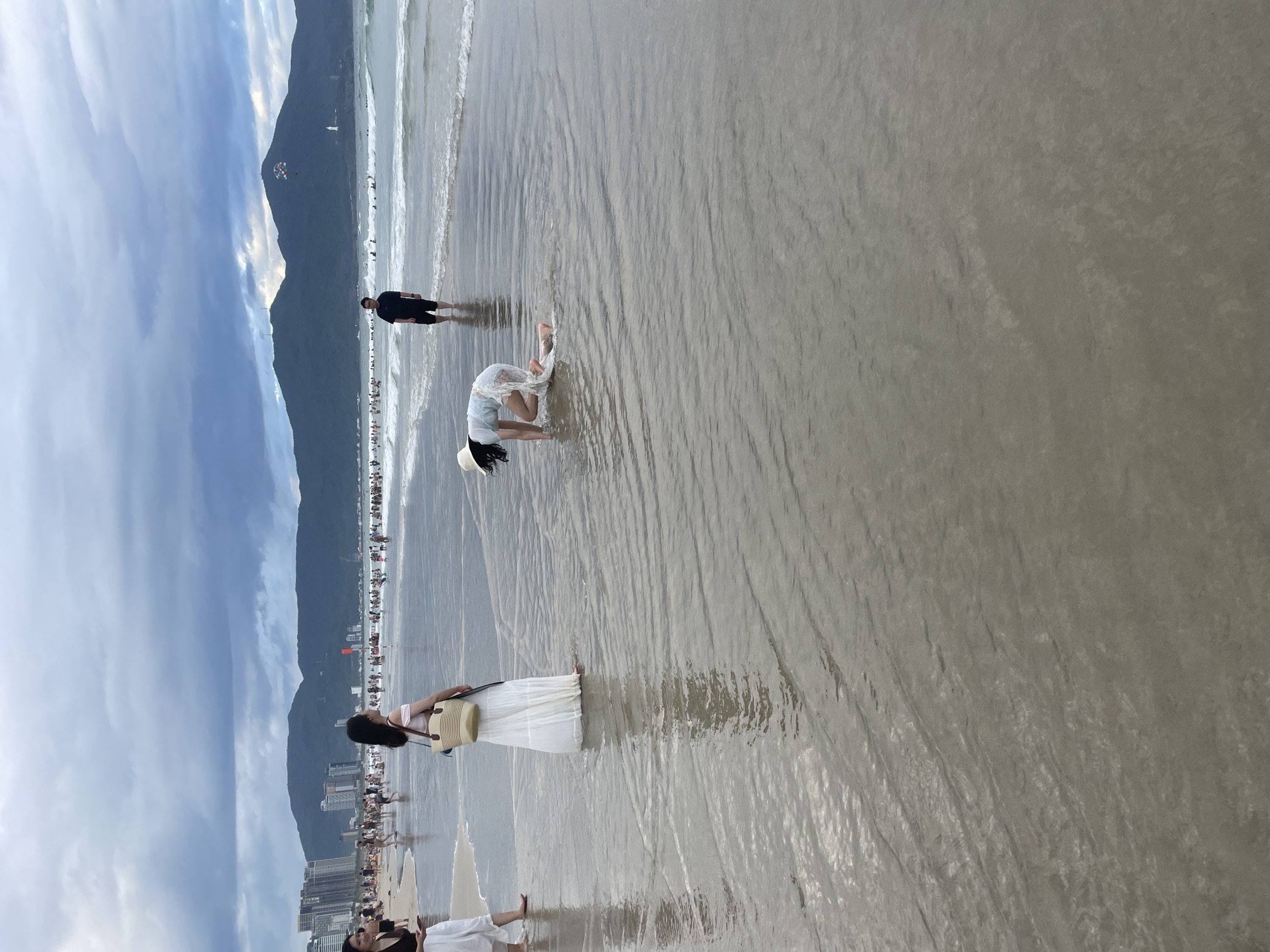 In Đà Nẵng, Vietnam, this image captures a scene at a beach, possibly My Khe Beach, under a cloudy sky. In the foreground, a woman is crouched down in the shallow water, her hat tilted forward as she appears to be interacting with the water. To her left, a woman in a long white skirt and a light-colored top stands facing the water, holding a straw bag. Further to the left, another woman is partially visible, wearing white pants. In the middle ground, a man in a dark t-shirt and shorts stands in the water, facing the camera. The background reveals a populated beach with many people wading in the ocean and standing on the shore. Beyond the beach, a line of buildings and lush green mountains form the cityscape and natural backdrop. The ocean is calm, with small waves rolling in.