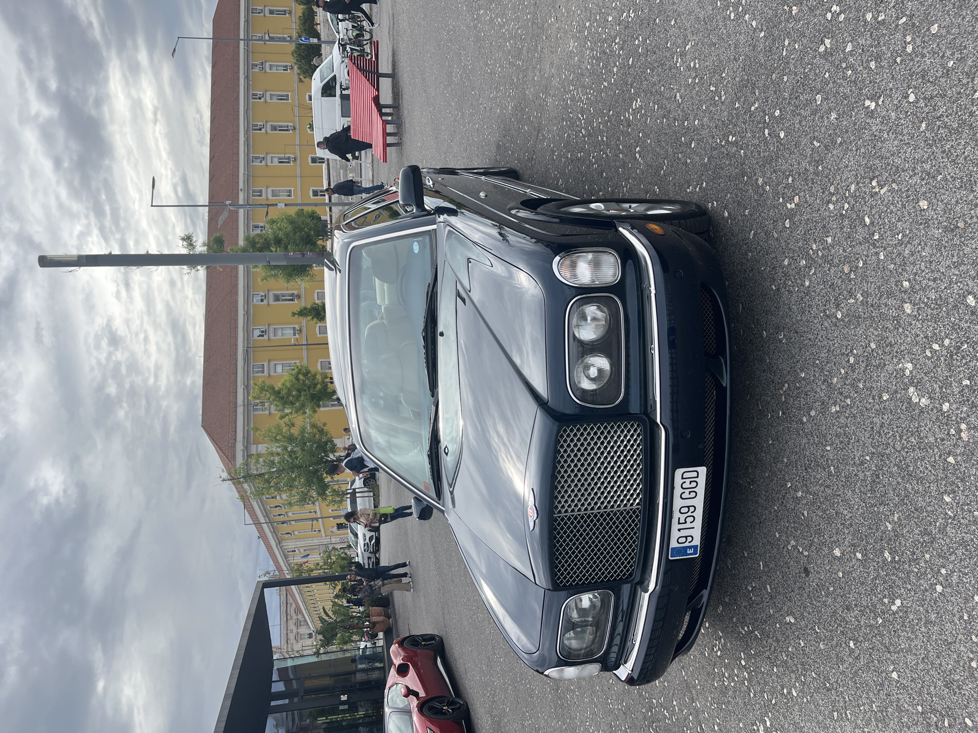 The image captures a dark blue Bentley Arnage prominently parked on a gray asphalt surface. The car's front grille, headlights, and European license plate ("9159 GGD") are clearly visible. Behind the car, a yellow multi-story building with numerous windows is a focal point, suggesting a public or institutional structure. In the mid-ground, there is a red bench and a white van, along with several people, most of whom appear to be walking or standing. The background includes portions of modern architecture, indicating a blend of old and new urban elements. The cloudy sky suggests an overcast day in Lisbon, Portugal, given the car's license plate which includes the European "E."