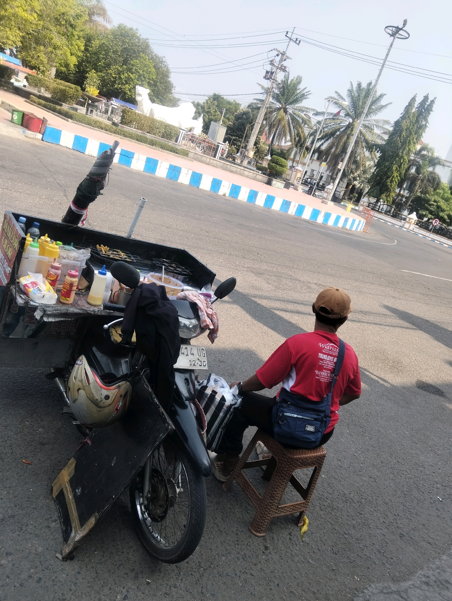 The image depicts a street scene in Ponorogo, Indonesia, where a vendor is selling food from a mobile cart attached to a motorcycle. The vendor, wearing a red shirt and a brown cap, is seated on a plastic stool, facing away from the camera. The motorcycle is parked on the asphalt road, with a black cart filled with food items and condiments. The cart has a visible sign with text, including "MAKLOR CILOR" and "MANDIRI MUSIMAN", suggesting the type of food being sold. Several bottles of sauces and tissues are arranged on the cart. A helmet rests on the front of the motorcycle, and a black fabric is draped over the handlebars. In the background, palm trees and other greenery are visible, along with streetlights and power lines, indicating an urban or semi-urban environment. A white statue of what appears to be a lion or tiger is also visible in the distance, possibly a landmark. The road is lined with blue and white striped barriers. The overall scene suggests a typical day for a street food vendor in Indonesia.