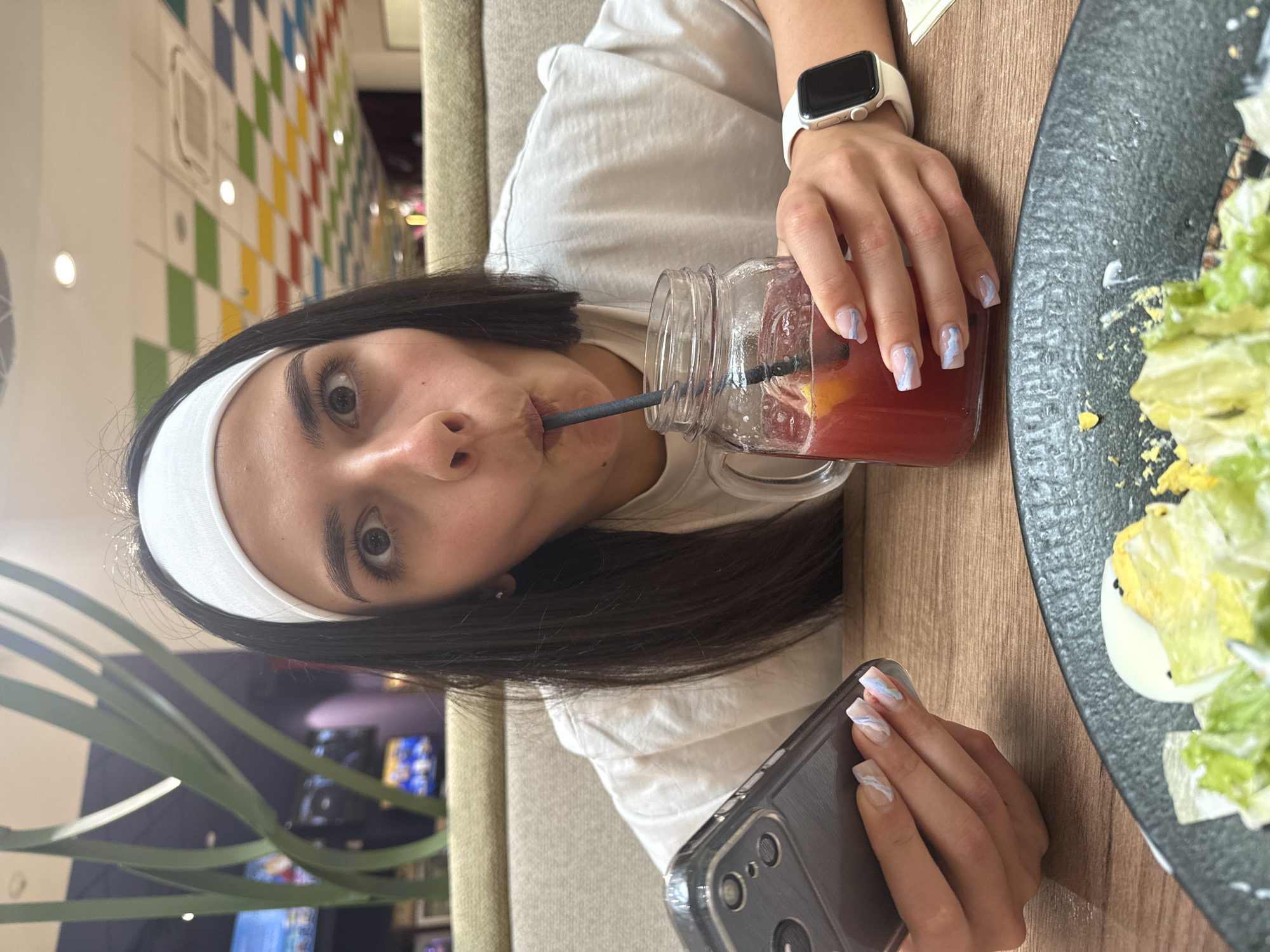 A young adult female, with long dark hair and a white headband, is depicted in a medium shot within an indoor commercial establishment in Veliko Tarnovo, Bulgaria. She is wearing a light-colored t-shirt and a white smartwatch on her left wrist. Her right hand holds a dark-colored smartphone in a clear case, while her left hand grasps a handled mason jar containing a red beverage with ice, a citrus slice, and a dark straw. She is actively drinking from the straw, with her eyes wide and lips pursed. Her fingernails are manicured with a light base and blue/white wavy design. On a wooden table in the foreground, a black plate partially displays a salad containing lettuce, hard-boiled egg, and a yellowish crumbled topping. The background features light beige upholstered booth seating. To the left, blurred green decorative elements and multiple television screens are visible. To the right, a ceiling with a multicolored checkered pattern (green, blue, yellow, red squares), recessed lighting, and an air conditioning vent is present.