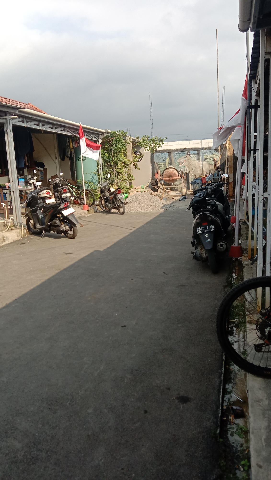 The image depicts a narrow, paved alleyway in Karangmangu, Indonesia, with buildings on either side. The scene is populated by several parked motorcycles, suggesting a residential or small business area. A bicycle wheel is visible in the foreground on the right, partially obscuring the view of the alley. In the background, there are two Indonesian flags displayed, one red and white, and the other red and white with stripes. The buildings feature simple structures with corrugated metal roofs and some open-air covered areas. The sky is overcast with clouds. The overall impression is one of a typical urban environment in Indonesia, possibly a residential neighborhood with some commercial activity.