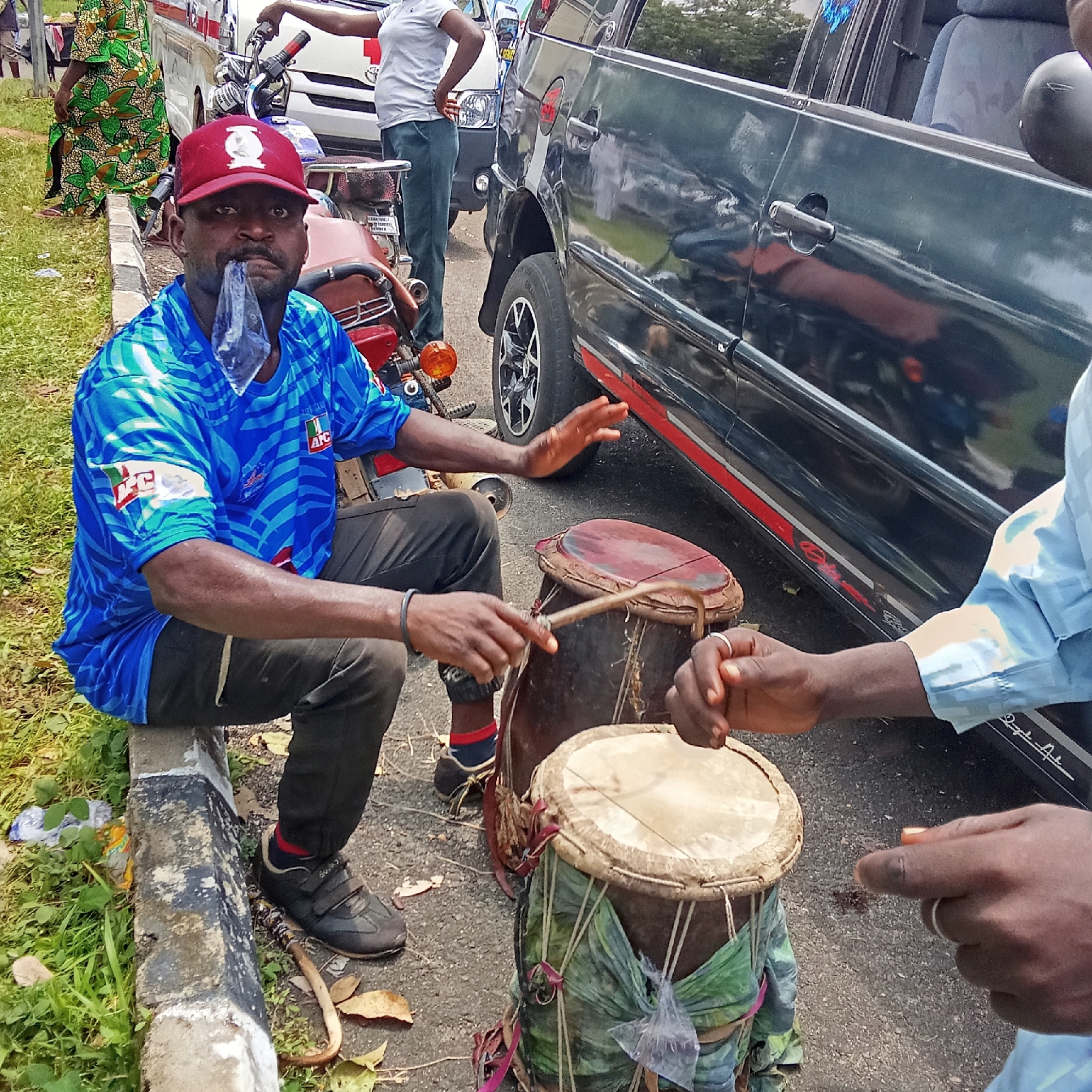 An African man in a blue and red baseball cap sits on a curb in Akwanga, Nigeria. He wears a blue shirt with a political logo and black pants. He is playing two drums with a wooden stick, while holding a clear plastic bag in his mouth. Another person’s hand is visible near the drums. In the background is a black vehicle, a motorcycle, and other people. The setting appears to be a street or roadside.