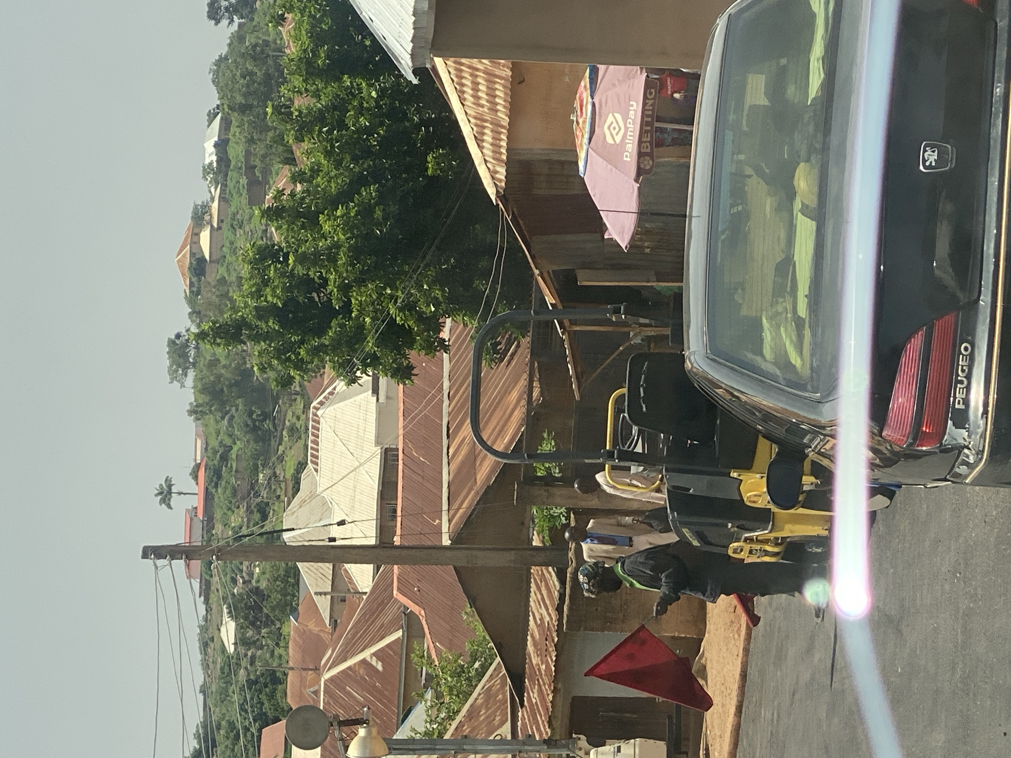 The image depicts a street scene in Jos, Nigeria, oriented vertically. In the foreground, a black Peugeot car is parked, its rear visible. The car's bumper has the word "PEUGEOT" partially visible. Behind the car, a yellow vehicle, possibly a small construction machine or a utility vehicle, is present. A person wearing a dark jacket and a patterned head covering is seen next to the yellow vehicle, appearing to be engaged in some activity. A red triangular flag is also visible on the ground.
The background shows a densely populated residential area with numerous buildings, many featuring corrugated iron roofs. The buildings are situated on a hillside, which is covered in lush green trees. A palm tree is visible on the left side of the hillside. The sky is overcast, suggesting it might be a cloudy day. The overall impression is one of a bustling, somewhat informal urban environment.
On a building to the left of the car, a purple umbrella with logos is visible. Text on the umbrella includes "PalmPay" and "BETTING". Overhead, telephone wires are strung between utility poles, indicating the presence of infrastructure. The time of day appears to be daytime, given the ambient light. There are no overt signs of distress or celebration, suggesting a normal daily scene.