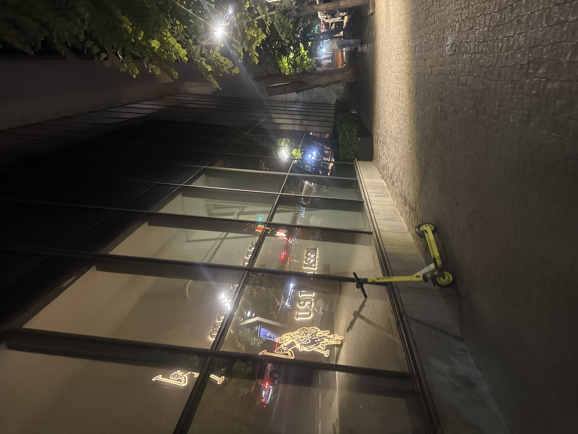 The image depicts a nighttime scene on a street in Tbilisi, Georgia, likely in a commercial or business district. The foreground shows a cobblestone sidewalk leading to the entrance of a building with large glass windows. A yellow electric scooter is parked on the sidewalk in front of the building. The glass windows of the building reflect the streetlights and traffic, as well as illuminated signage. Inside the building, a neon sign featuring a polo player on a horse is visible, along with the text "US POLO ASSN. 1890." In the background, trees line the street, and more streetlights illuminate the scene. There are a few cars and streetlights visible in the reflections and on the street. A person is walking away from the camera on the sidewalk in the middle ground to the right of the frame.
