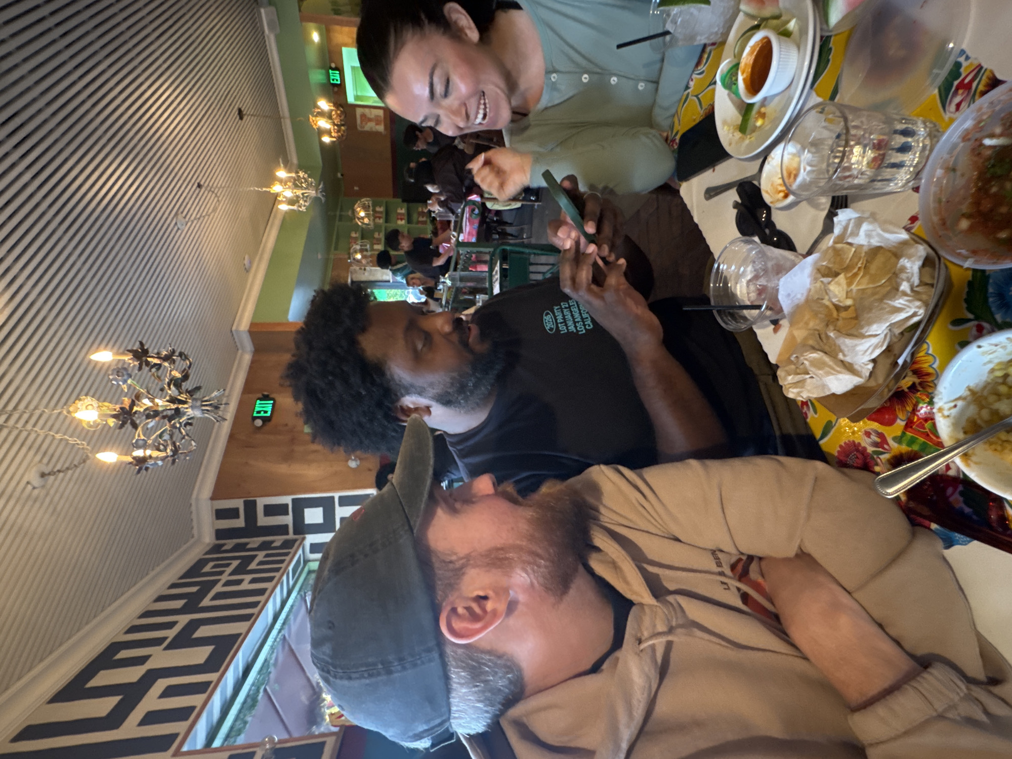 Three individuals are captured at a table setting, likely in a restaurant or eating establishment within the city of Los Angeles, California, USA. The woman to the right is fair-skinned and laughing while looking at the cell phone held by the person to the left of her. The man holding the phone has dark skin, a beard, and curly black hair. He is wearing a black t-shirt. To his left, a man with a light skin tone, beard, and wearing a baseball cap, is turned away from the camera and rests his arm on the table. The table is covered with a colorful patterned cloth and has several dishes and glasses on it, suggesting that the people are finishing their meal. There are chips, salsa, and other food remnants on the table. In the background, a section of the restaurant is visible, showing other patrons at tables, decorative chandeliers hanging from the ceiling, and architectural details like striped walls and an exit sign.