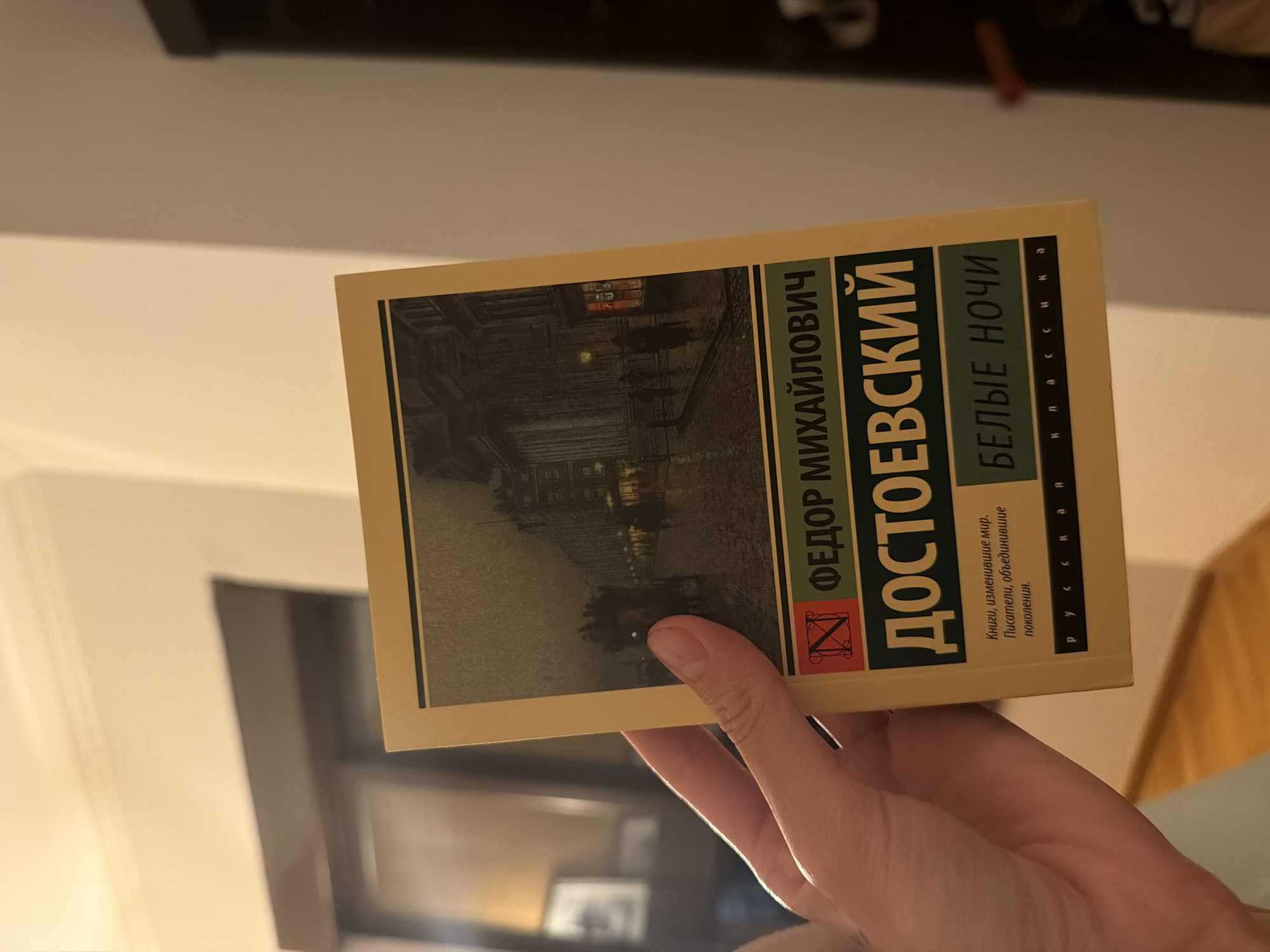 A hand holds a yellow book titled "Белые ночи" (White Nights) by ФЕДОР МИХАЙЛОВИЧ ДОСТОЕВСКИЙ (Fyodor Mikhailovich Dostoevsky). The book's cover features a dark, atmospheric illustration of a street scene at night with a carriage and figures walking. The background of the image is out of focus, showing a dimly lit room with a window and a dark piece of furniture.