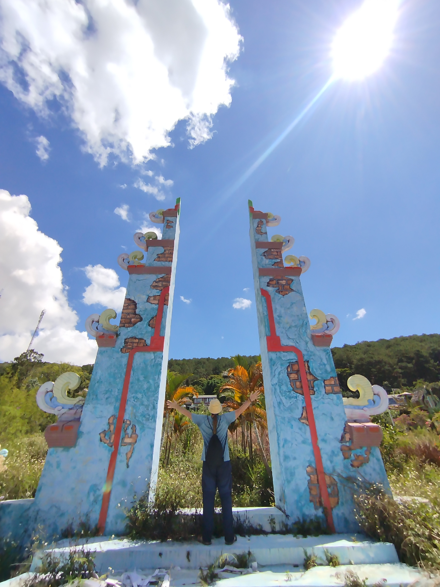 A low-angle, wide shot depicts a person standing within a decaying, colorful archway under a bright sky with scattered clouds and strong sunlight. The archway, comprised of two blue pillars decorated with faux brickwork and stylized ornamentation, appears weathered and neglected. The person, facing away from the camera with outstretched arms, is wearing a light-colored hat, a blue shirt, and dark pants. The surrounding area is overgrown with weeds and sparse vegetation. In the background, a wooded hillside is visible. The scene is located in the city of Da Lat, Vietnam.