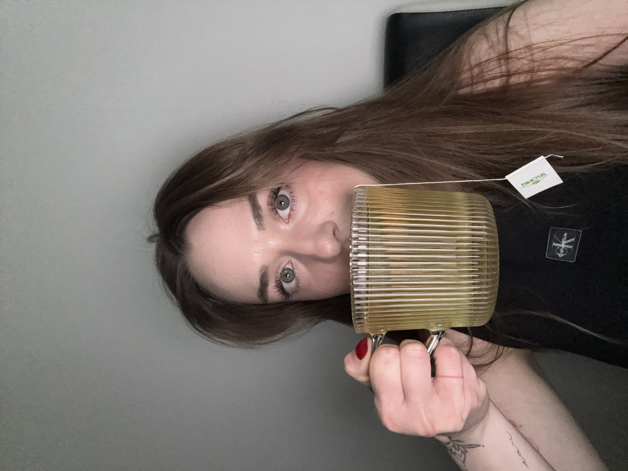 A woman with long brown hair holds a gold ribbed mug with a tea bag inside, positioned in front of her face. She has fair skin, green eyes, and manicured fingernails painted red. She is wearing a black Calvin Klein t-shirt. The background consists of a gray wall and a black chair. No specific location in Kragujevac, Serbia can be determined from the image.