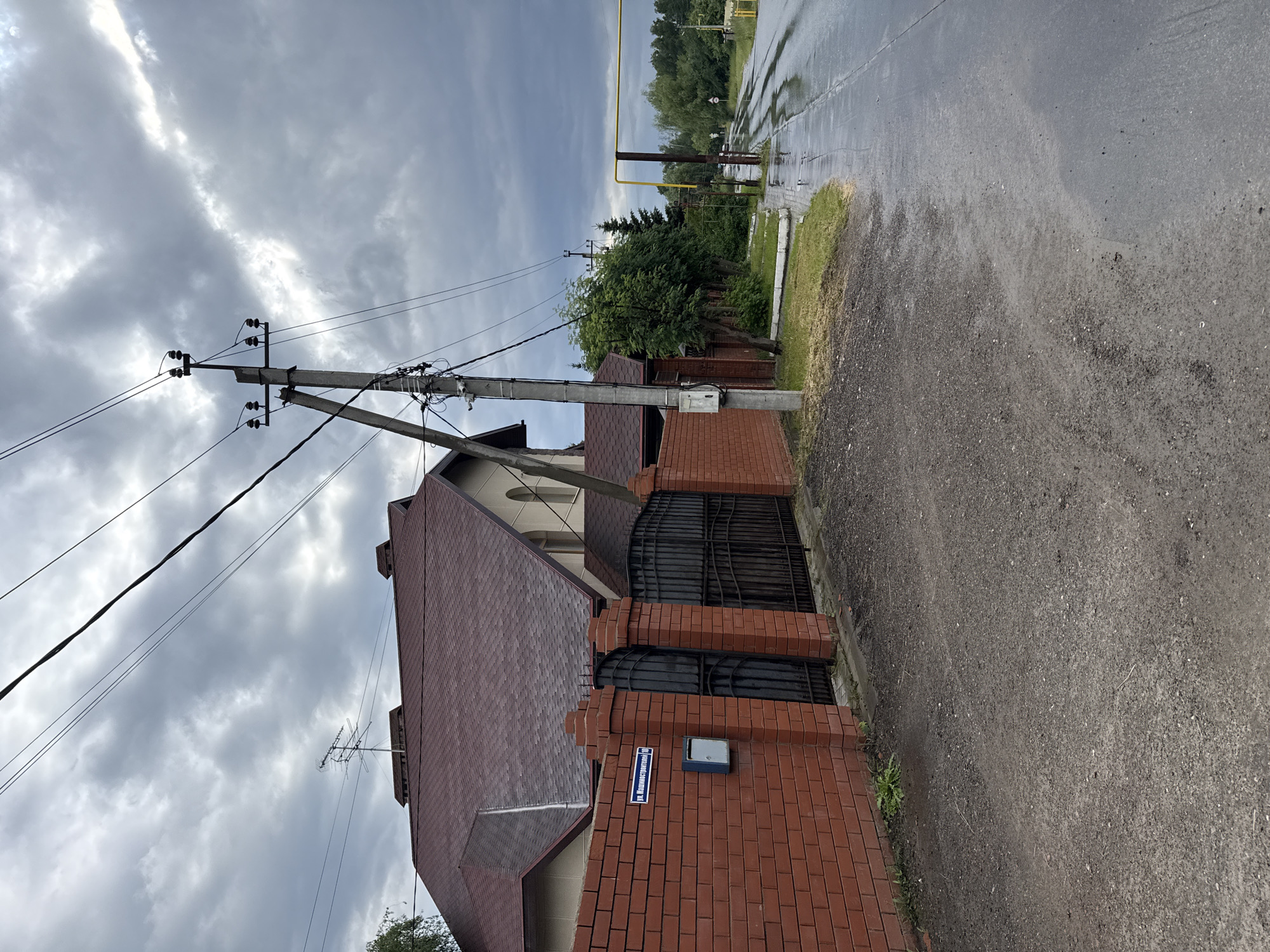 The image shows a street scene in Kazan, Russia, under a cloudy sky. A two-story house with a dark red, shingled roof and brick exterior is prominent. A tall brick fence with a black metal gate surrounds the property. A street sign on the fence reads "ул. Ямашева" (Yamasheva Street).
To the left of the house, a concrete utility pole with numerous wires is visible, carrying electricity to the buildings. A gas pipeline runs parallel to the street in the background. The asphalt road appears wet, suggesting recent rain. Dense green trees and foliage line the opposite side of the street.
The overall mood is somewhat somber due to the overcast weather, with heavy, gray clouds dominating the sky. The scene depicts a residential street with typical urban infrastructure. No people are present in the image. The time of day is difficult to determine precisely due to the lack of direct sunlight, but the overcast conditions suggest it could be daytime, possibly late afternoon or early evening.