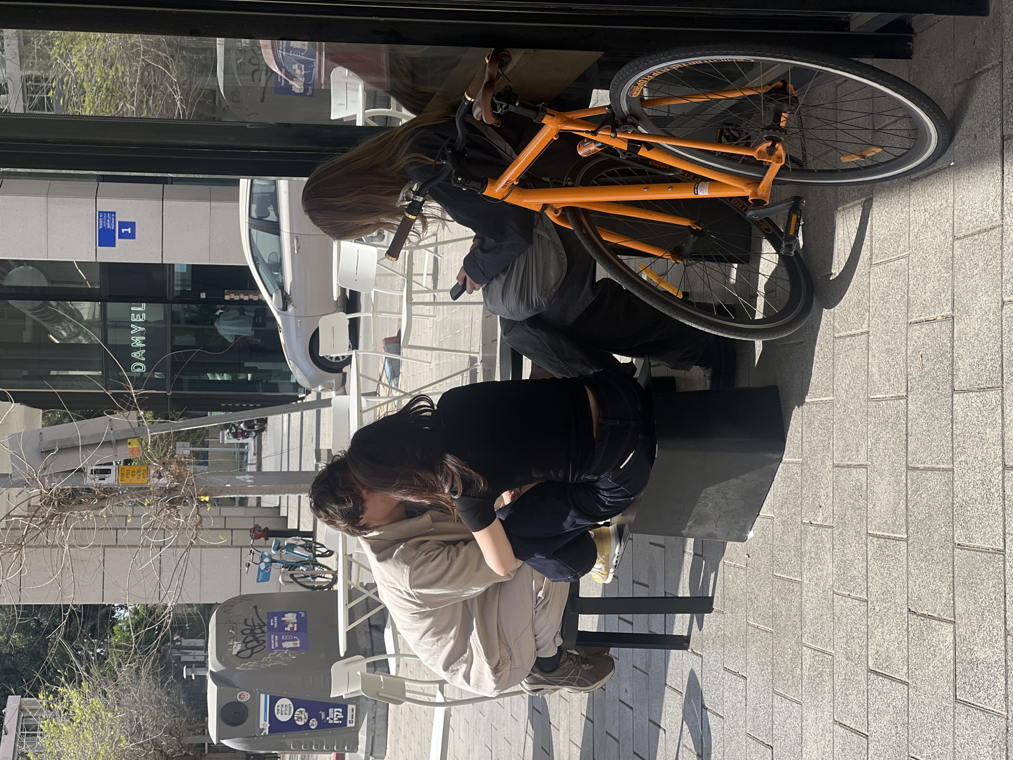 A view from inside a building looking out onto a Tel-Aviv street scene. Two individuals are embracing on a bench. One person is wearing a beige jacket and sneakers. The other individual is wearing a black shirt, dark pants, and yellowish sneakers. An orange bicycle rests against the window. Outside, a white van is visible, along with a street sign written in Hebrew. In the background, there is a trash can with graffiti and posters on it and a bicycle rack with blue bikes. The street is paved with light-colored stone. Bare branches are visible on the left side of the frame. A sign for the establishment "DANYEL" can be seen through the window.