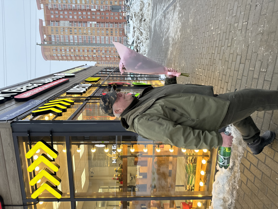 An adult male is observed walking from left to right along a brick-paved sidewalk at Ulitsa Triumfalnaya 11a, Odintsovo, Russia. The man wears a camouflage-patterned baseball cap, an olive-green hooded jacket, olive-green trousers, and dark boots. He carries a bouquet of flowers wrapped in pink paper in his right hand, tied with a pink ribbon. In his left hand, he holds a green plastic bottle with a white cap. The scene is set in front of a modern commercial building with large glass windows and black frames. Prominent signage on the building includes "Ул. Триумфальная 11а" and "ЦВЕТЫ" (Flowers). Bright yellow arrow-shaped decorations adorn the building's facade. Through the store windows, various plants and merchandise are visible. Patches of dirty snow line the base of the building and form larger piles in the distant background. Beyond the snow, parked vehicles and several tall, multi-story residential buildings with orange and light brown facades are visible under an overcast sky, indicating winter conditions.