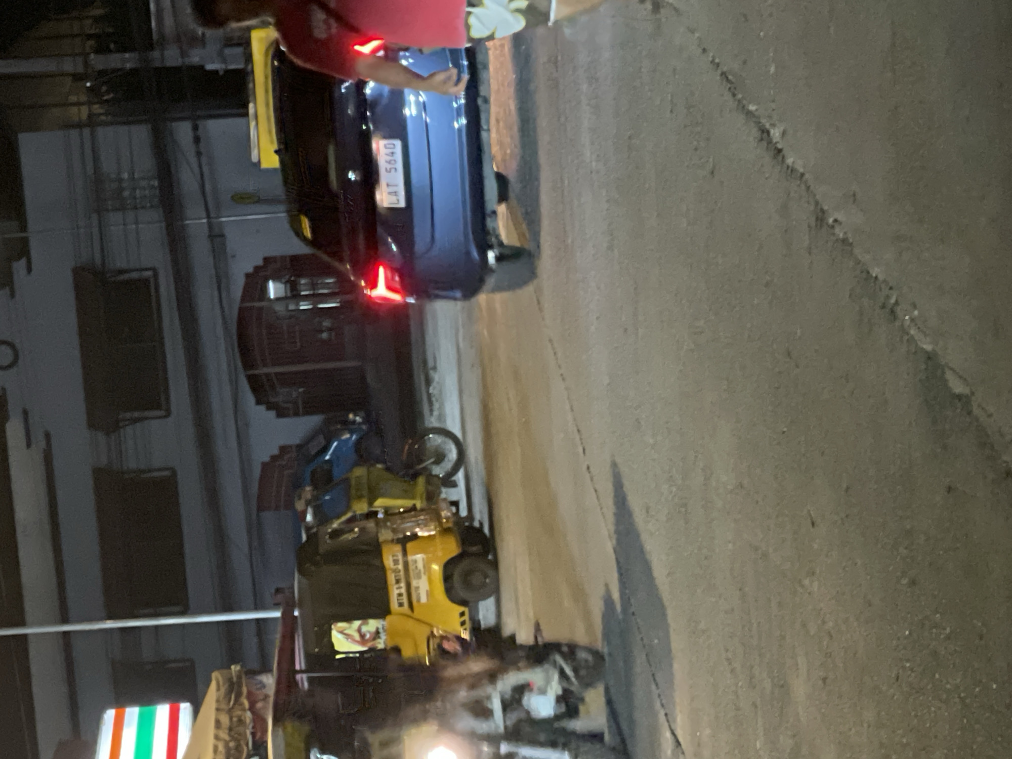 The image captures a nighttime scene on a city street, likely in Davao City, Philippines, given the presence of motorized tricycles commonly used for public transportation there. In the foreground, the rear of a dark blue sedan is visible, displaying a license plate that reads "LAT 5640". The car's brake lights are illuminated. To the left of the sedan, several yellow motorized tricycles are parked or moving slowly. One tricycle in the foreground is partially obscured and has its headlights on. A person wearing a red shirt and patterned shorts is seen on the right side of the frame, partially facing away from the camera. The environment appears to be an urban setting with buildings visible in the background, some of which have illuminated windows. The street surface is paved and shows signs of wear. The overall lighting suggests it is after dark, with artificial lights from vehicles and buildings illuminating the scene.