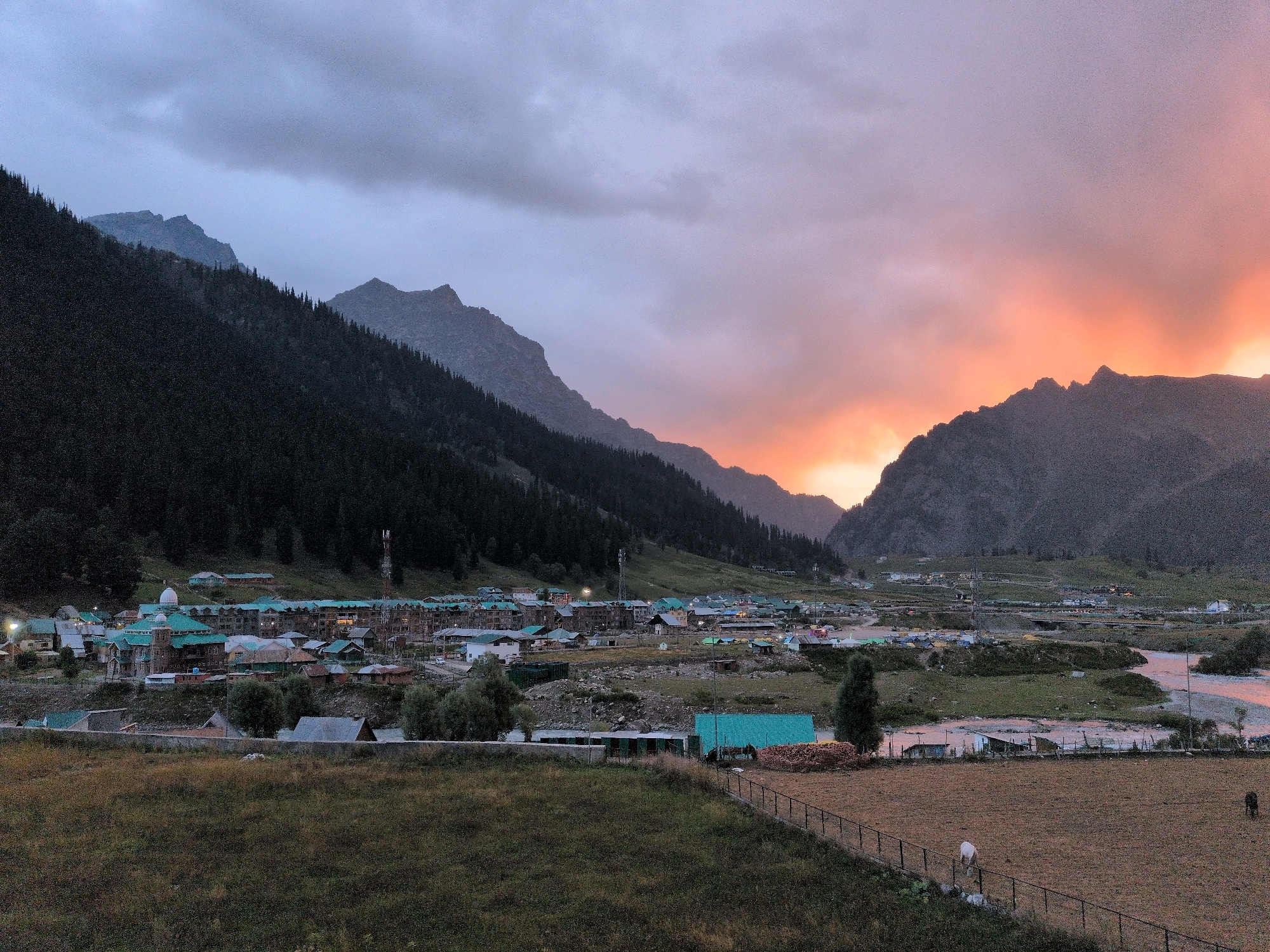 The image depicts a scenic landscape at dusk in Sonamarg, India, with a village nestled at the foot of a mountain range. The sky displays a gradient of colors from deep blue and gray clouds on the left to a vibrant orange and pink sunset on the right. The mountains are covered with dark green pine trees on the lower slopes, becoming more barren and rocky towards the peaks.
In the mid-ground, a village is visible with numerous buildings featuring green roofs. Some buildings appear to be clustered together, forming a town center, while others are more spread out. A prominent white dome structure stands out among the buildings. Light poles with illuminated lamps are scattered throughout the village, indicating artificial lighting. A communication tower is also visible on a hill behind the village.
The foreground consists of a grassy field with some patches of dry, brown grass. A black metal fence runs horizontally across the image, separating the grassy field from a cultivated area. Within the fenced area, a white horse grazes on the right side, and a dark brown cow is visible further to the right. A small river or stream flows through the middle ground, reflecting the colors of the sky. The overall impression is one of a peaceful, rural setting at the end of the day, with the natural beauty of the mountains and the warm hues of the sunset dominating the scene.
