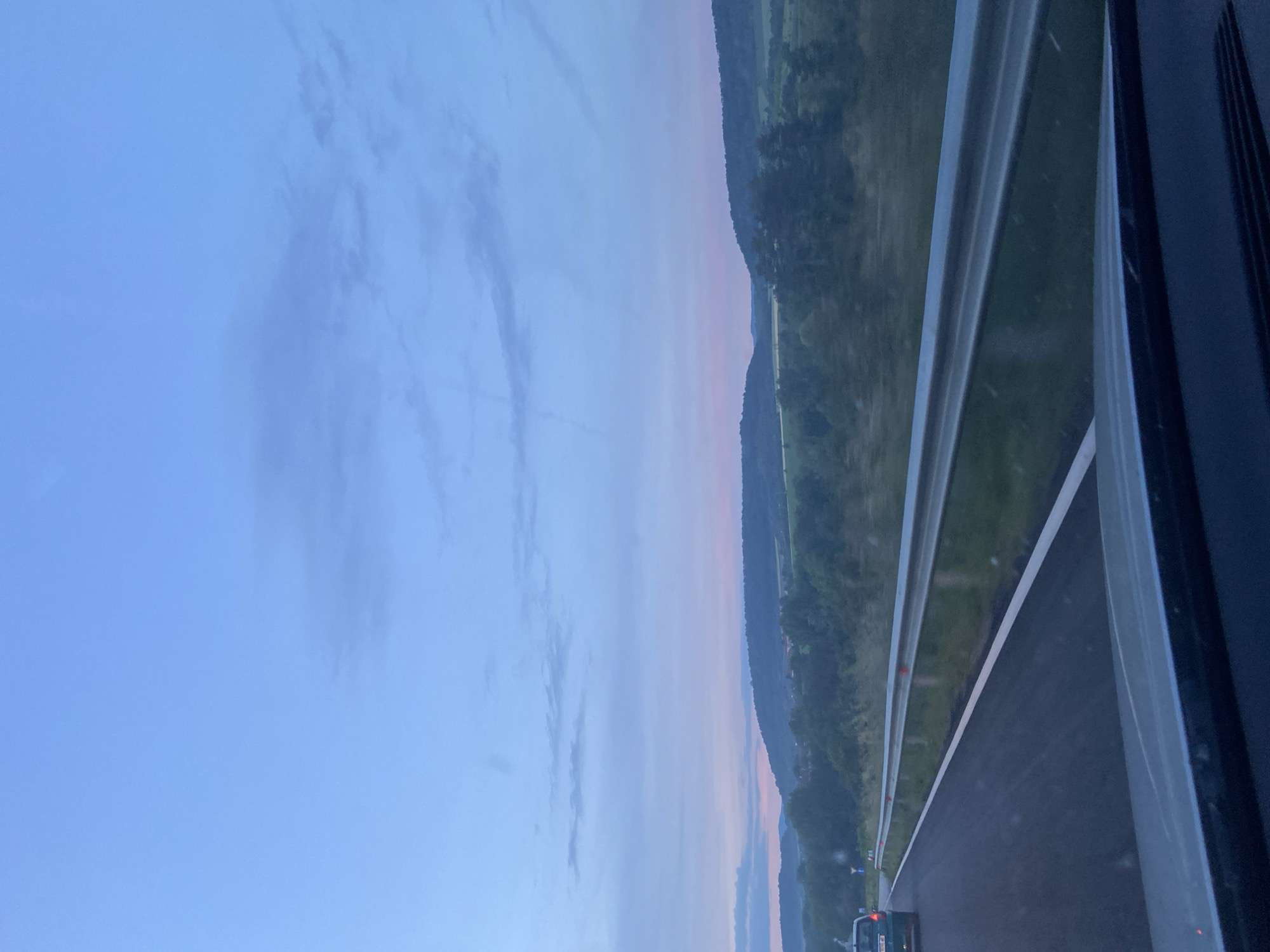 The image depicts a highway landscape, likely near Dragijevo, Bulgaria, based on the GPS coordinates. It seems to be taken from inside a moving vehicle, evident from the side of the car and the motion blur. To the left, the sky is a pale blue with some clouds. In the center, the highway stretches out, bordered by a green, hilly landscape. The lighting suggests it might be early morning or late evening, indicated by the soft light in the sky. A vehicle is visible on the road ahead.