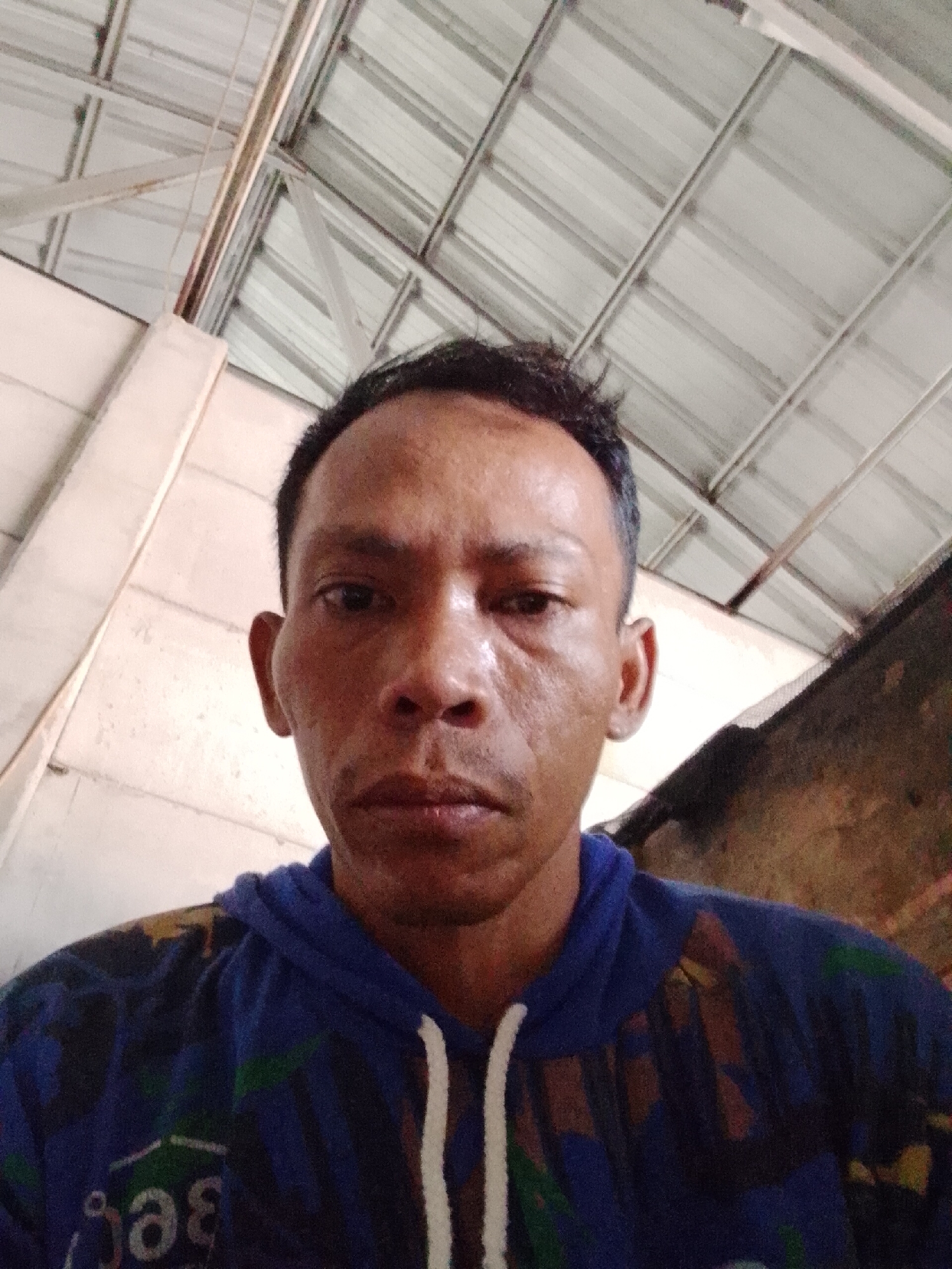 A man takes a selfie in Bukit Ketok, Indonesia. The man has dark, short hair and is wearing a blue hooded sweatshirt with white drawstrings. The background shows the interior of a building with white walls and a metal ceiling.