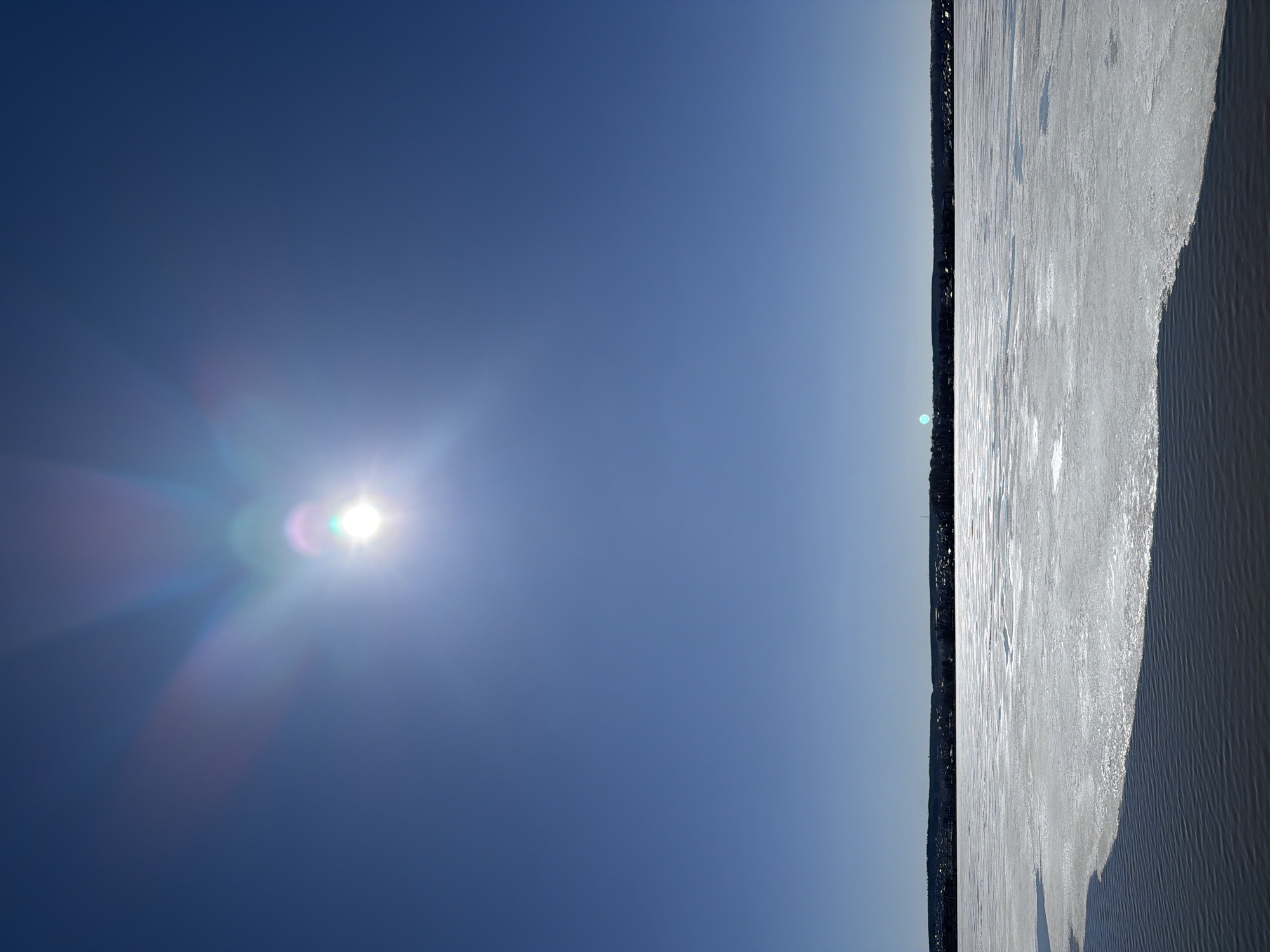 The image presents a winter scene in Votkinsk, Russia. The sky is a clear, bright blue with the sun prominently visible on the left side, creating a lens flare effect. A narrow strip of land is barely visible on the horizon, marking the edge between the sky and a frozen body of water, possibly the Votkinsk Reservoir. The ice surface is a pale, reflective white, exhibiting varying textures and possibly thin layers of snow. The right side of the image shows dark water near the bank. The overall scene evokes a sense of cold and clarity.