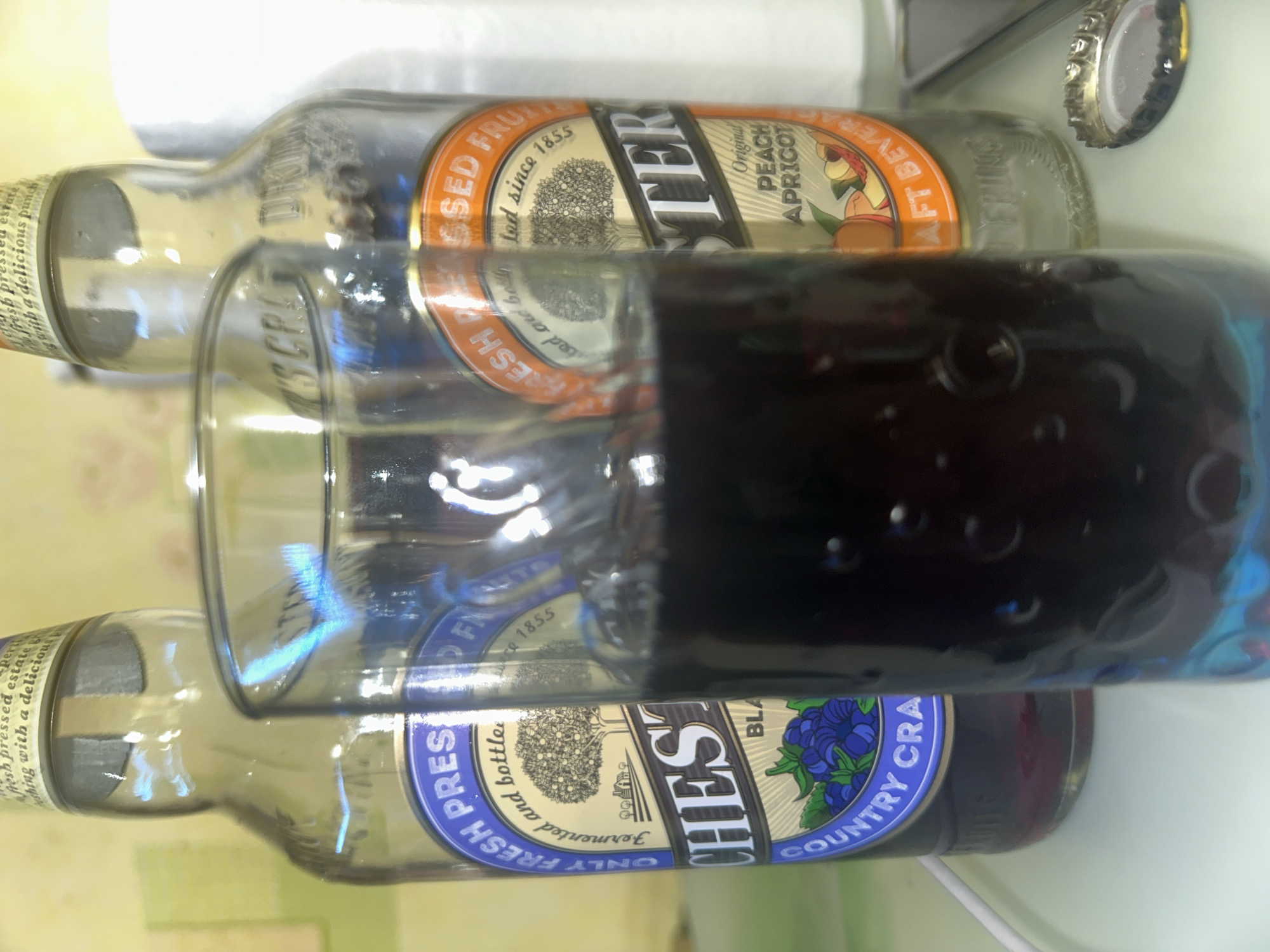 The image shows two bottles of 'Chester's' brand beverages lying on their sides. One is 'Original Peach Apricot' flavor, the other 'Country Craft Blackberry'. A glass filled with dark liquid is placed between the bottles, partially obscuring them. The scene suggests a casual, indoor setting. A bottle cap is visible in the top right corner of the frame. The OCR text suggests the bottles claim "fresh pressed estate" ingredients, fermented, and bottled since 1855. There is no information in the image suggesting a location in Moscow, Russia.