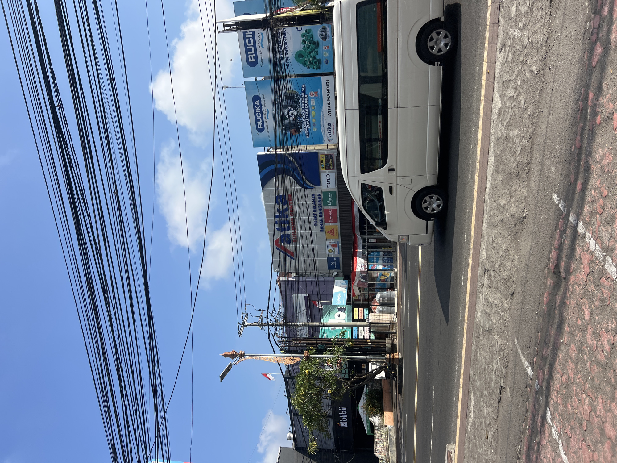 In Denpasar, Indonesia, on a clear day with a bright blue sky and scattered clouds, a white passenger van drives down a street lined with buildings and overhead power lines. The van is positioned in the right lane of a multi-lane road. To the left of the street, a sidewalk runs in front of a row of businesses. One building displays a large sign with the word "Atika" and "BUILDING CENTER" in blue lettering. Below this sign, there are smaller signs advertising various brands. Another building to the far left features the "blibli" logo. A street lamp with ornate detailing stands on the sidewalk, adorned with a small Indonesian flag. Trees and bushes are visible along the street. The street itself has yellow lane markings and a visible curb.