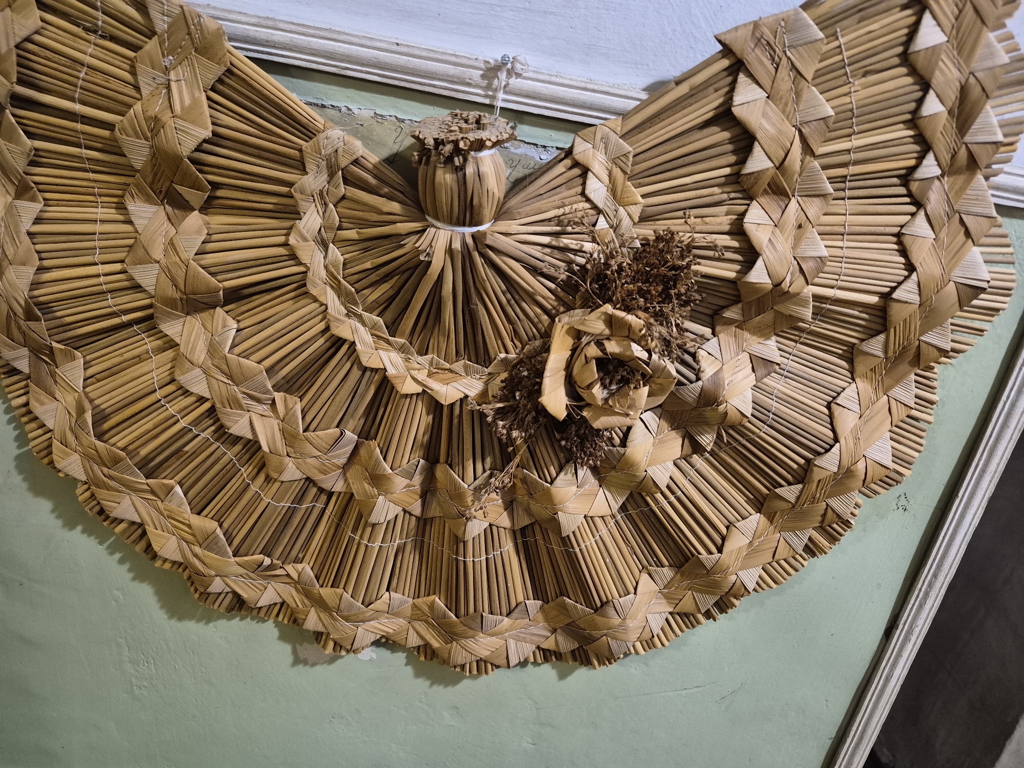 An ornate, fan-shaped wall decoration crafted from dried straw is displayed on a pale green wall in Vanadzor, Armenia. The decoration features intricate weaving techniques forming a tiered, semi-circular pattern that radiates outwards from a central element resembling a doll or angel. This central figure is formed from straw tubes, with a small, cylindrical base topped by a textured cap, and is bound by a white string. To the right of the central figure, a decorative element made of dried brown leaves and a straw rose adds further texture. The straw decoration is attached to the wall by a thin white string tied to a small nail or hook above it. The wall exhibits signs of age with some paint wear and faint, almost illegible writing visible in places. A white, ornate molding runs along the top of the wall where it meets the ceiling. The lighting casts shadows on the wall, suggesting it is an indoor scene.