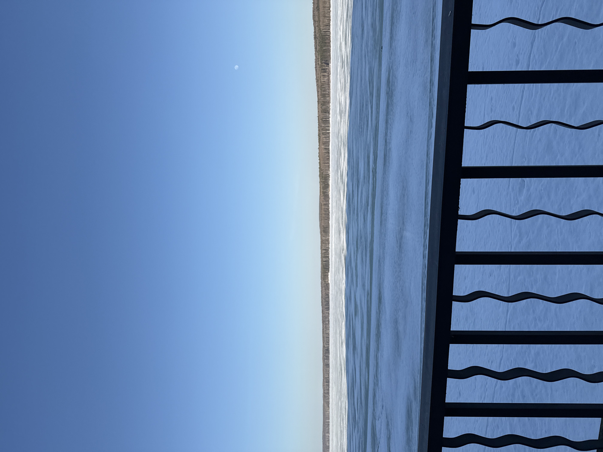The image shows a body of water, possibly the Kama River, with a shoreline of trees in the distance. The sky is blue with a small, pale circle visible in the upper left. A dark metal structure with wavy bars runs vertically along the right side of the image. The perspective suggests the photograph was taken from a high vantage point. The location is likely near the city of Chaikovsky, Russia.