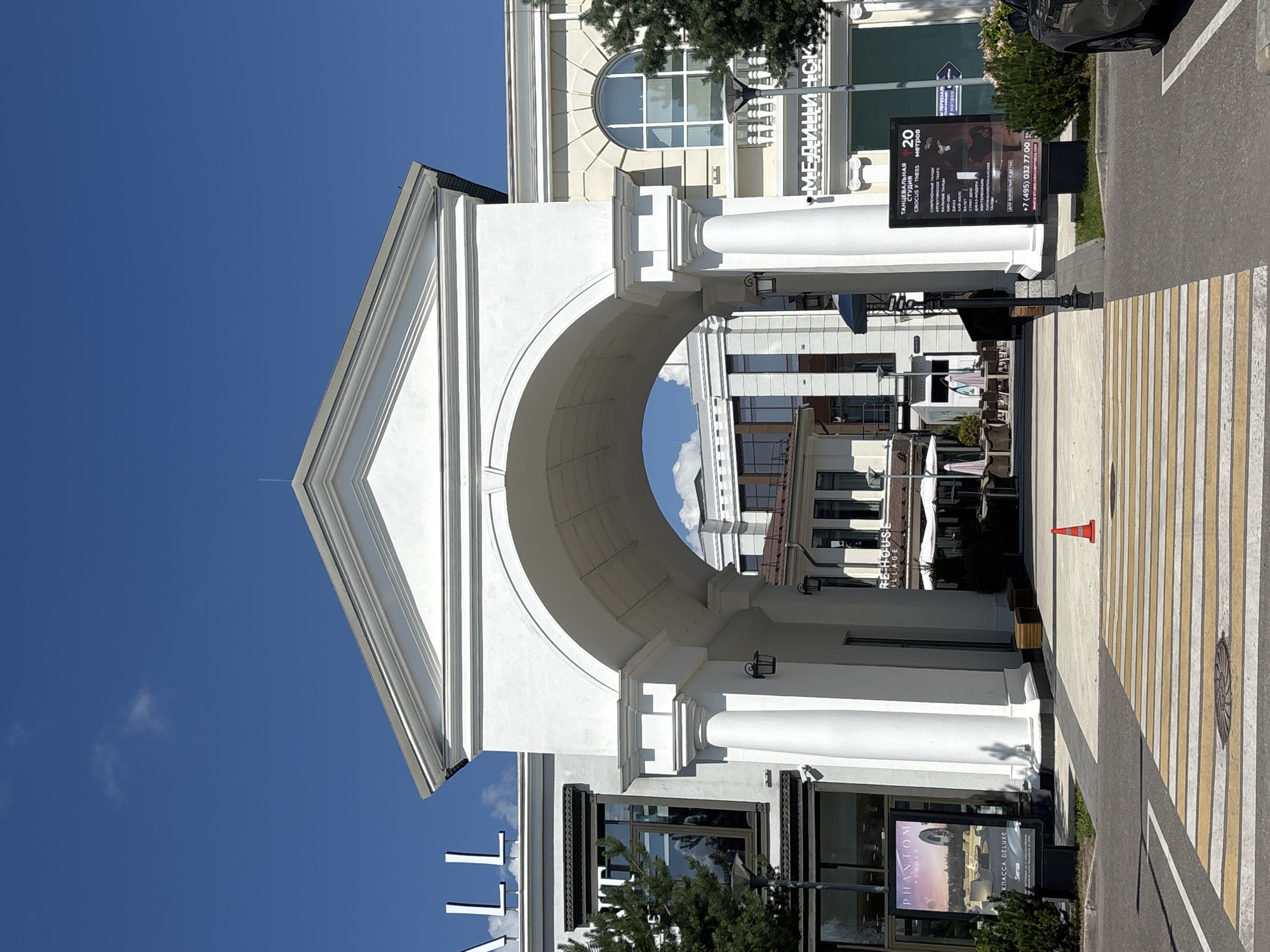 The image depicts an outdoor scene in the city of Millennium Park, Russia. In the foreground, a pedestrian crosswalk with yellow and white stripes is visible. An orange traffic cone is placed on the crosswalk to the right of center. Behind the crosswalk, a large white archway with columns leads into a courtyard area. On the left side of the archway, a billboard advertises "PHANTOM" and "KLASSADELUXE". On the right side of the archway, another billboard advertises a dance studio, "TANTSEVALNAYA STUIDIYA CROCUS F TNESS", with class listings and contact information.
Beyond the archway, the courtyard features buildings with windows and awnings, some with outdoor seating. Trees and planters are present in the area. The sky is clear and blue with a few wispy clouds. The architecture of the buildings suggests a modern, possibly commercial or residential complex. The overall impression is a bright, sunny day in a well-maintained urban space.
