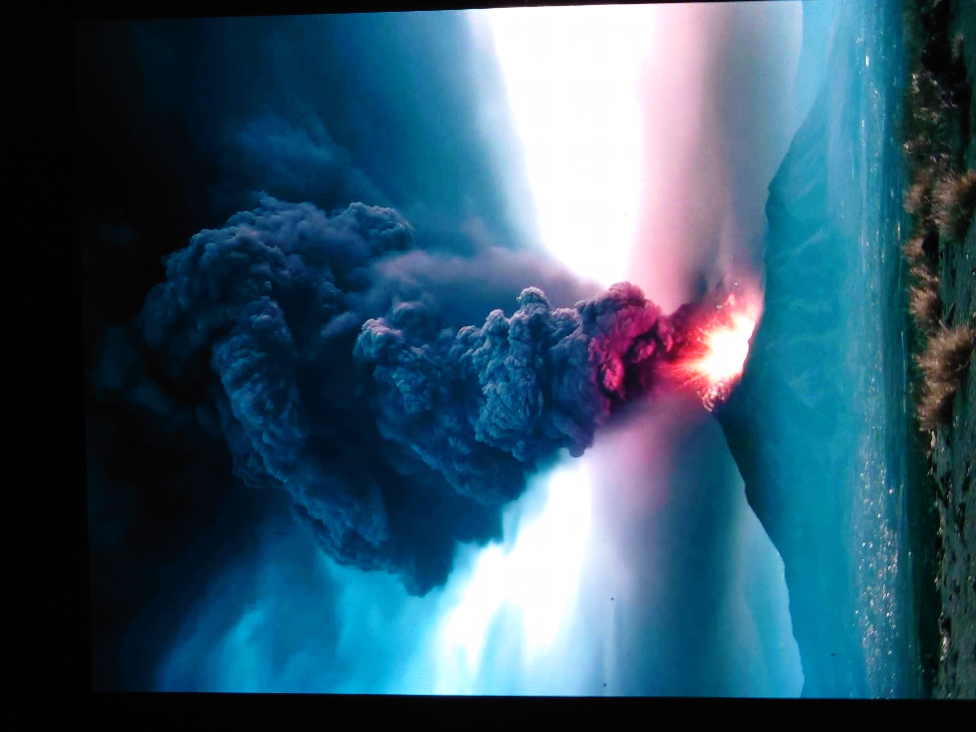 A dramatic volcanic eruption dominates the scene. A massive plume of dark ash and smoke billows upwards from the summit of a cone-shaped mountain. The eruption is intensely bright at the peak, with fiery red and pink hues emanating from the crater and casting a glow on the surrounding smoke. The sky is a mix of dark, ominous clouds and areas of bright, hazy light, possibly from the volcanic glow or atmospheric conditions. In the distance, a city or town is visible, its lights twinkling faintly through the haze. The foreground consists of a dark, rocky landscape with sparse, dry vegetation. The image captures the raw power and destructive potential of a volcanic event. The mention of Belorechensk, Russia, is not supported by any visual evidence in the provided image.