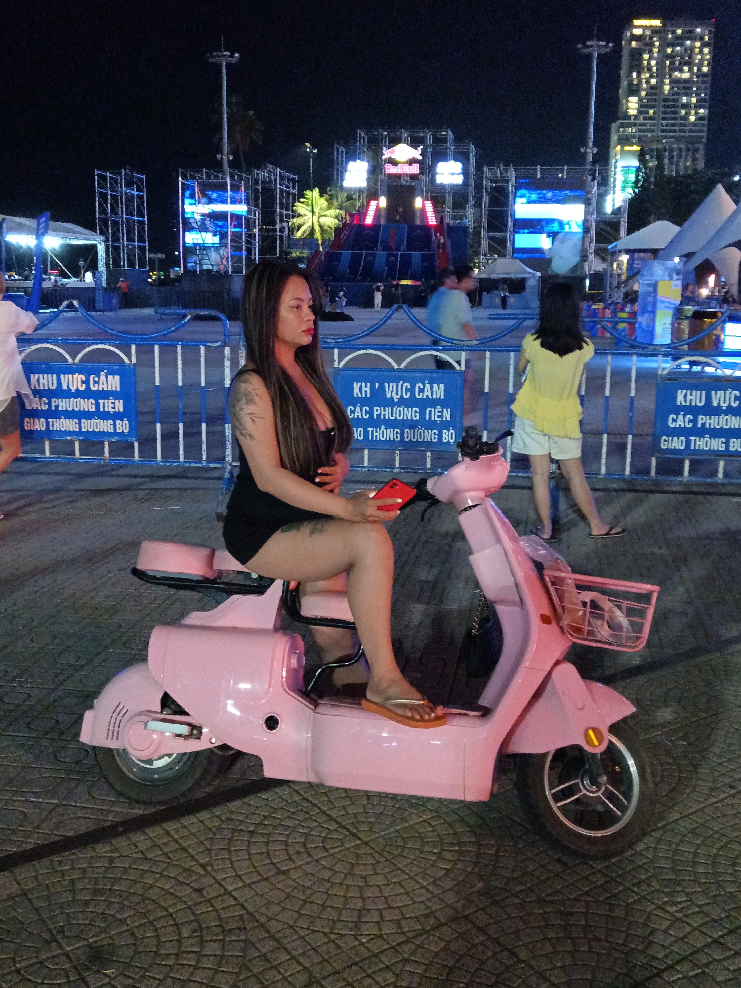 The image depicts a woman sitting on a pink electric scooter at night, possibly at a street event or festival. She is facing to her right, with her legs crossed, and has tattoos on her arms and thigh. The scooter has a basket on the front and a black bag hanging from the handlebars. In the background, there are illuminated stage structures with large screens, palm trees, and tall buildings, suggesting an urban setting. Blue fencing is visible in the foreground, with signs in Vietnamese that translate to "No Parking Zone" and "Traffic Ban Zone". The ground is paved with textured tiles. The overall lighting suggests artificial illumination from the event and surrounding buildings.