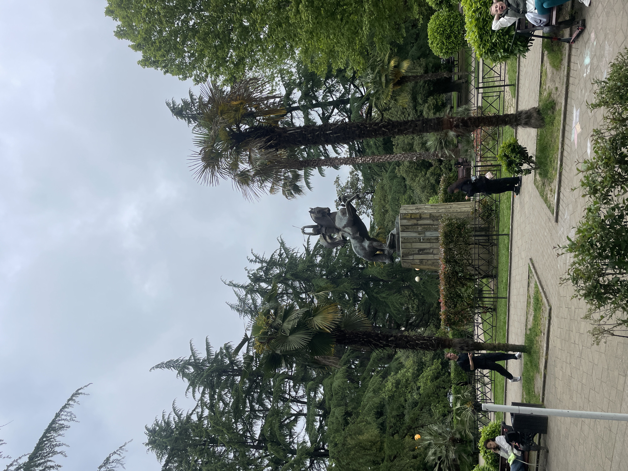 The image shows a statue of a horse and rider in a park setting. The statue stands atop a stone base and is surrounded by lush greenery, including palm trees. A paved walkway runs along the right side of the image, with people sitting on benches and walking along the path. The sky is overcast. The location is Sochi, Russia.