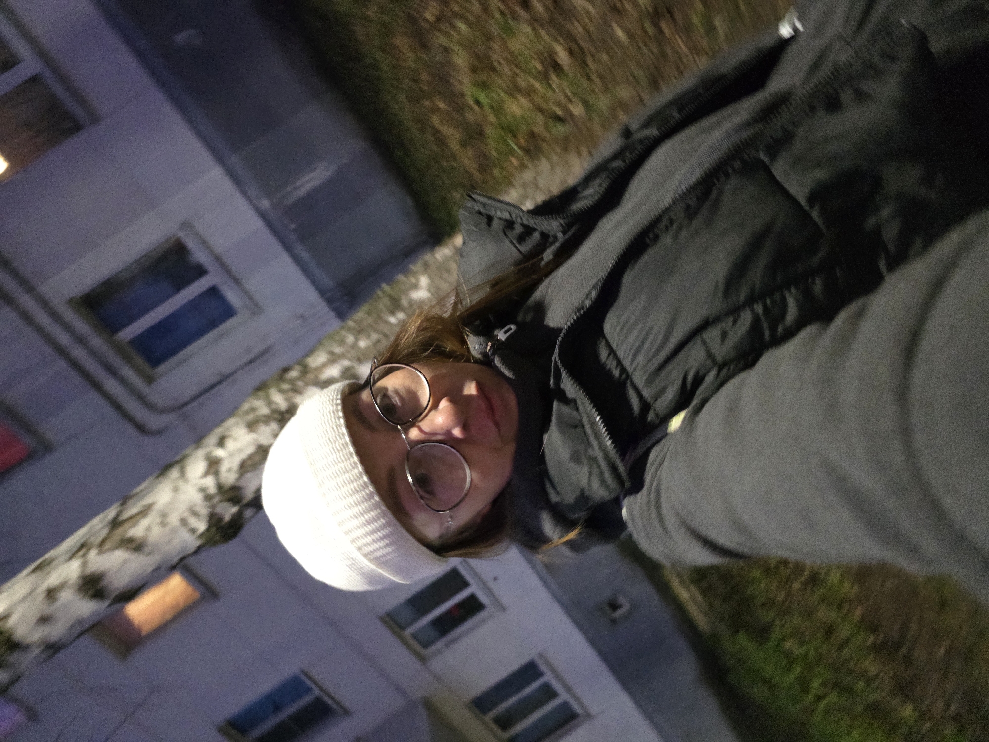 A woman wearing a white beanie and round glasses is facing the camera. She is wearing a dark puffer jacket and a grey sweatshirt. Behind her is a multi-story building with illuminated windows and a birch tree. Grass is visible in the upper right corner. The photo seems to be taken in a city at night. Given that the location is Chaikovsky, Russia, this is likely a residential or urban area in that city.