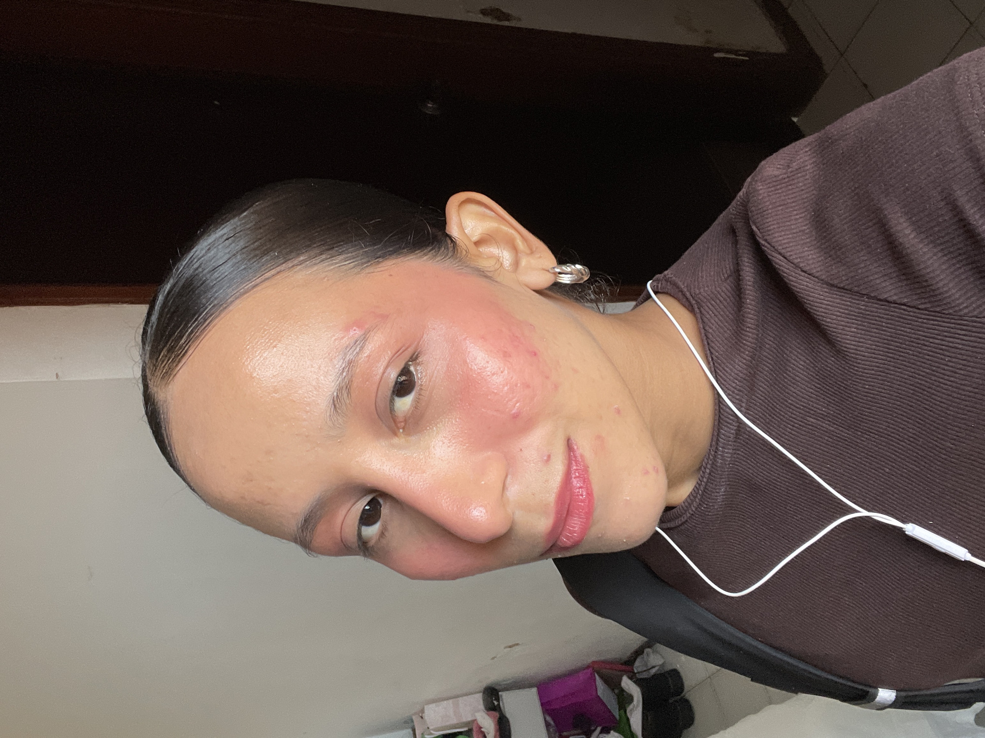 A woman with dark hair pulled back is taking a selfie. She is wearing a brown ribbed top and has earbuds in. Her face is flushed with pink blush, and she has some acne on her cheeks and chin. She is also wearing hoop earrings. The background appears to be an indoor setting with a door and part of a room visible. The image is a close-up of her face and upper body. The location is identified as Porlamar, Venezuela, although there are no specific visual cues in the image to confirm this.