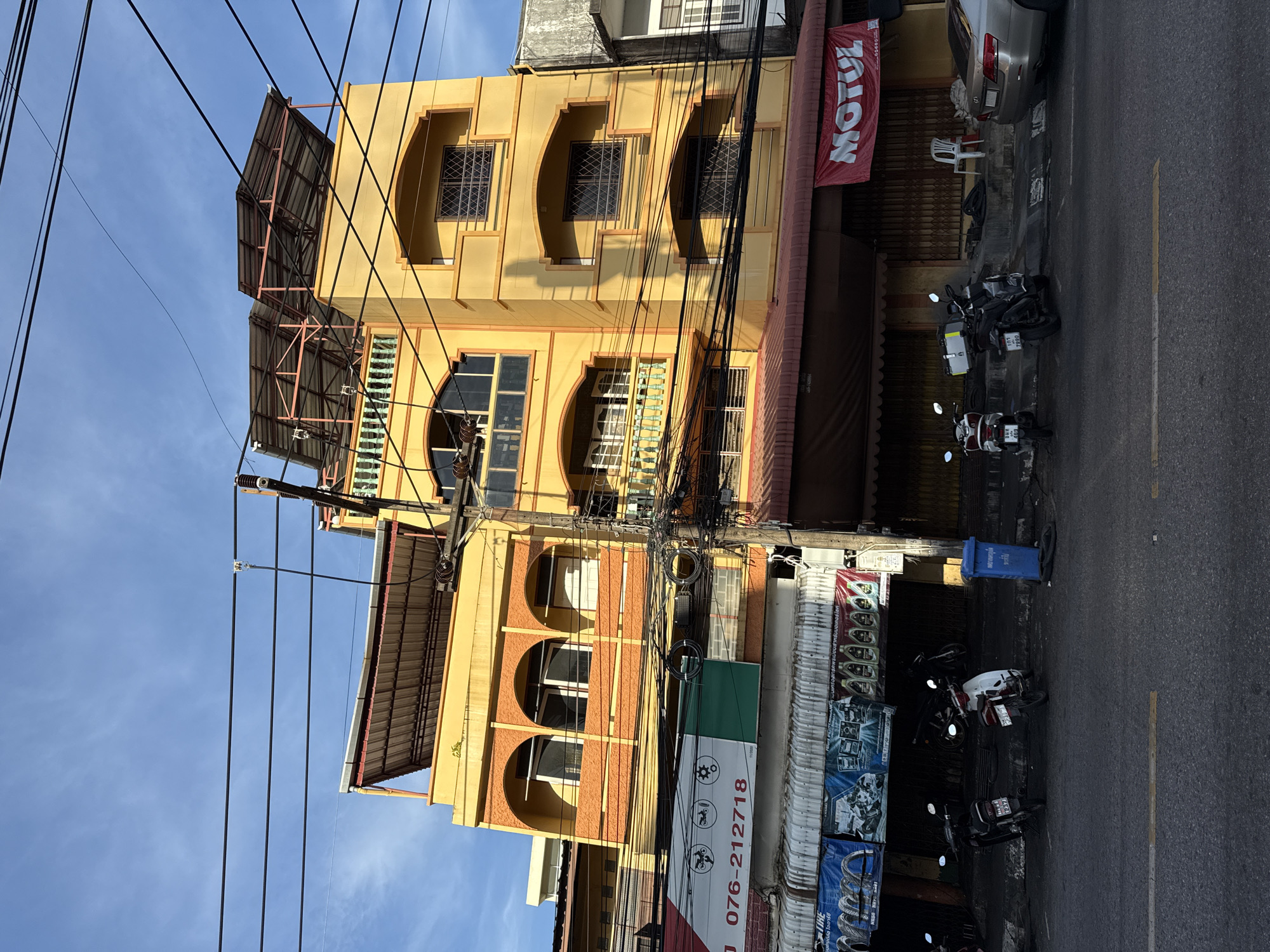 This is a medium shot, low-angle, street-level photograph taken in Phuket City Municipality, Thailand, showing a commercial building on the left and a street with parked motorcycles on the right. The building is a multi-story structure with a predominantly yellow facade, accented with orange arches and trim. The ground floor appears to be a commercial establishment with a red awning and roll-up shutters. Above the awning, there are various signs, including one with the text "MOTUL" in red letters. Several motorcycles are parked along the sidewalk in front of the building. Electrical wires are extensively strung across the foreground, obscuring parts of the building and the sky. The sky is blue with some light clouds. The overall scene depicts a typical urban streetscape in Southeast Asia, characterized by dense electrical infrastructure and commercial activity.