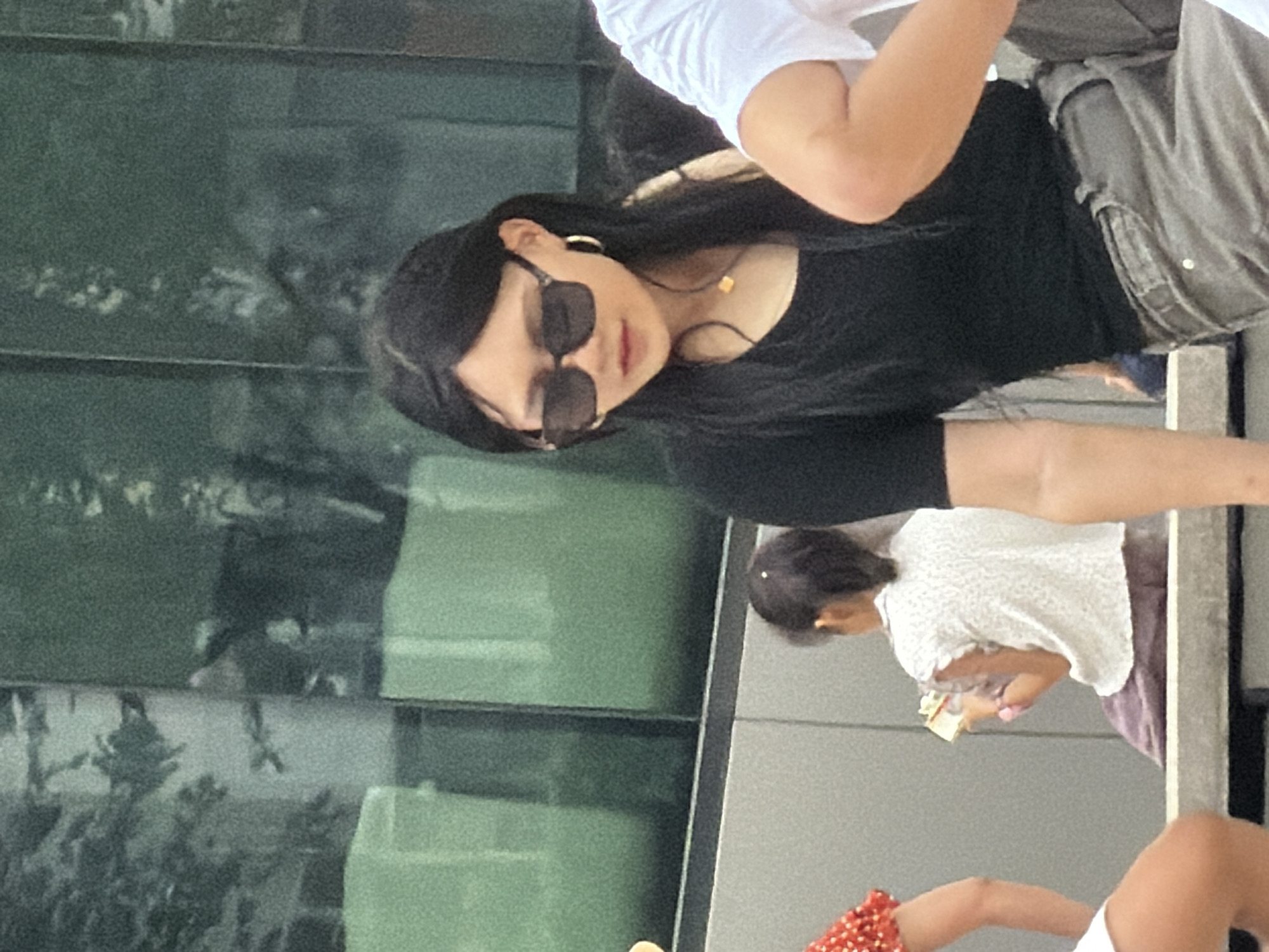 A medium shot captures a young woman wearing a black t-shirt, grey pants, and sunglasses, seated in the foreground. She has long, dark hair and is wearing a gold necklace and hoop earrings. To her left, a young child in a white shirt and purple skirt is visible, holding a small object. To the right, parts of two other individuals' arms are in the frame, one wearing a white t-shirt and the other a black t-shirt. The background consists of a green, reflective building facade and a grey concrete wall. The scene appears to be in an outdoor public space within Yuzhong District, China.
