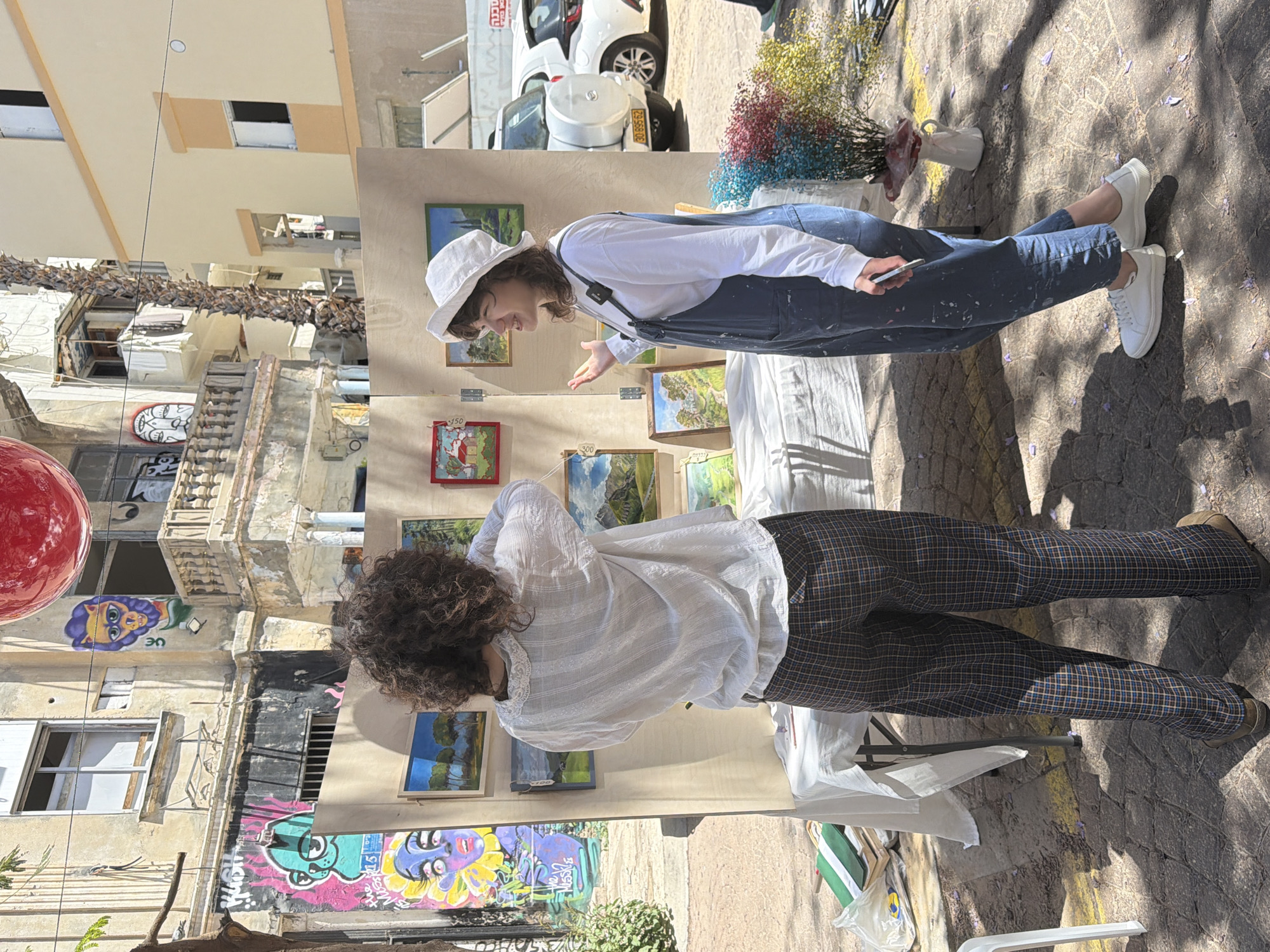 The image captures an outdoor art fair, likely in Tel Aviv, Israel, given the urban architectural style and presence of Hebrew text on a vehicle. Two individuals are at the center. The first person, wearing a white hat and denim overalls, faces an art display and extends a hand, possibly discussing a piece with the second person. This individual, with curly hair, wears a white shirt and plaid pants and is looking at the art display as well. A series of small paintings hang on the display. Various urban elements surround the scene, including graffiti-covered walls, buildings, and a parked white car in the background. Flowers in a white vase are located near one of the individuals.