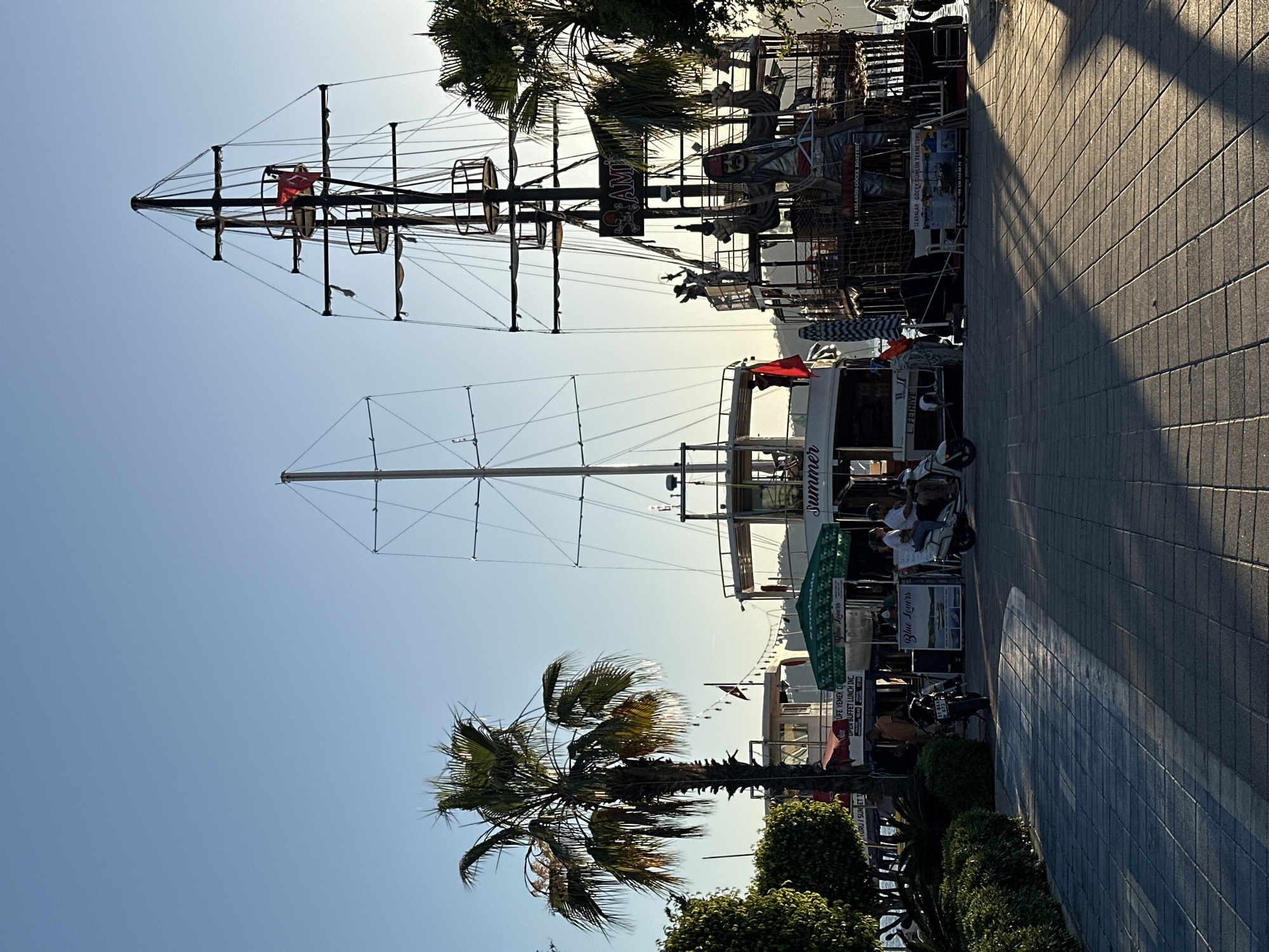 The image displays a sunny waterfront scene in Fethiye, Turkey, likely captured during the morning or late afternoon given the bright, directional sunlight casting long shadows across the paved promenade. The setting is a bustling dockside area popular with tourists.
A large, multi-masted tourist boat resembling a pirate ship dominates the left side of the frame, featuring intricate rigging, a red flag with a white emblem, and decorative pirate figures. Further along the dock, another boat bears the name "Summer" and "L. FETHIYE" on its side. Several people are visible, some sitting at tables under green "Blue Lovers" umbrellas, while one person is seen riding a scooter on the promenade.
Advertising signs for boat tours are prominent, with text such as "12 ADALAR GÖCEK GÜNLÜK TEKNE TURU" (12 Islands Göcek Daily Boat Tour) and "OPEN BUFFET LUNCH INC." Palm trees and lush green bushes line the promenade, adding to the leisurely atmosphere. The sky is clear and blue, indicating pleasant weather, ideal for the boat excursions suggested by the numerous vessels and signage.