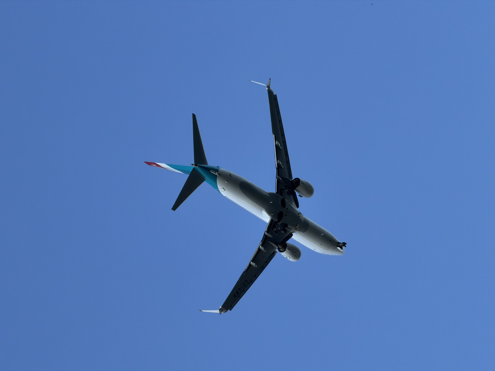 An aerial photograph captures an airplane flying against a clear, bright blue sky. The underside of the aircraft is visible, showing its wings extended, engines mounted, and landing gear deployed. The tail of the airplane features a distinctive red, white, and teal design. Visible on the wing is the registration number "LX-LBA". The perspective suggests the airplane is ascending or descending, viewed from directly below. The photograph was taken over Burgas, Bulgaria, indicated by the presence of the registration LX-LBA which is associated with Luxembourg Airlines, and Burgas being a major hub for air traffic in Bulgaria, often with international flights.
