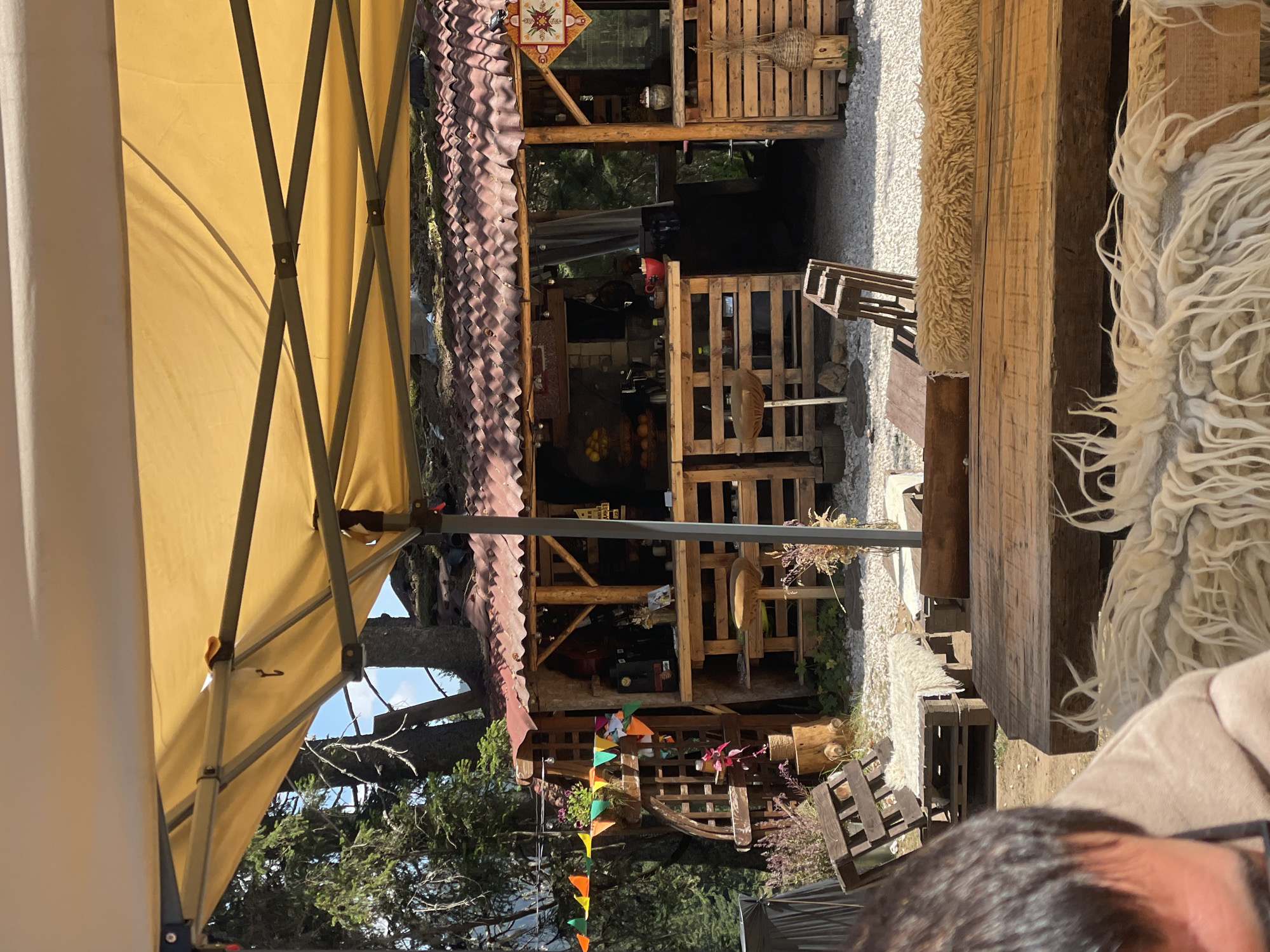 The image depicts an outdoor seating area of a rustic establishment, likely a cafe or bar, situated in a mountainous, forested environment. A large yellow canopy shades the foreground. In the background, a wooden structure with a corrugated metal roof serves as a counter and bar area. The bar is stocked with bottles, a red bowl, and what appears to be fruit in a bowl. Pallets are repurposed as shelving and seating. Decorative elements include colorful triangular flags strung across the front of the bar area and a woven decorative object on the right side of the bar. The ground is covered in light-colored gravel. A wooden table and seating, adorned with shaggy, off-white throws, are visible in the foreground. A person's head and shoulders, out of focus, are visible in the lower left corner of the frame. The overall atmosphere is casual and natural, fitting for an outdoor venue in a natural setting near Smolyan, Bulgaria.