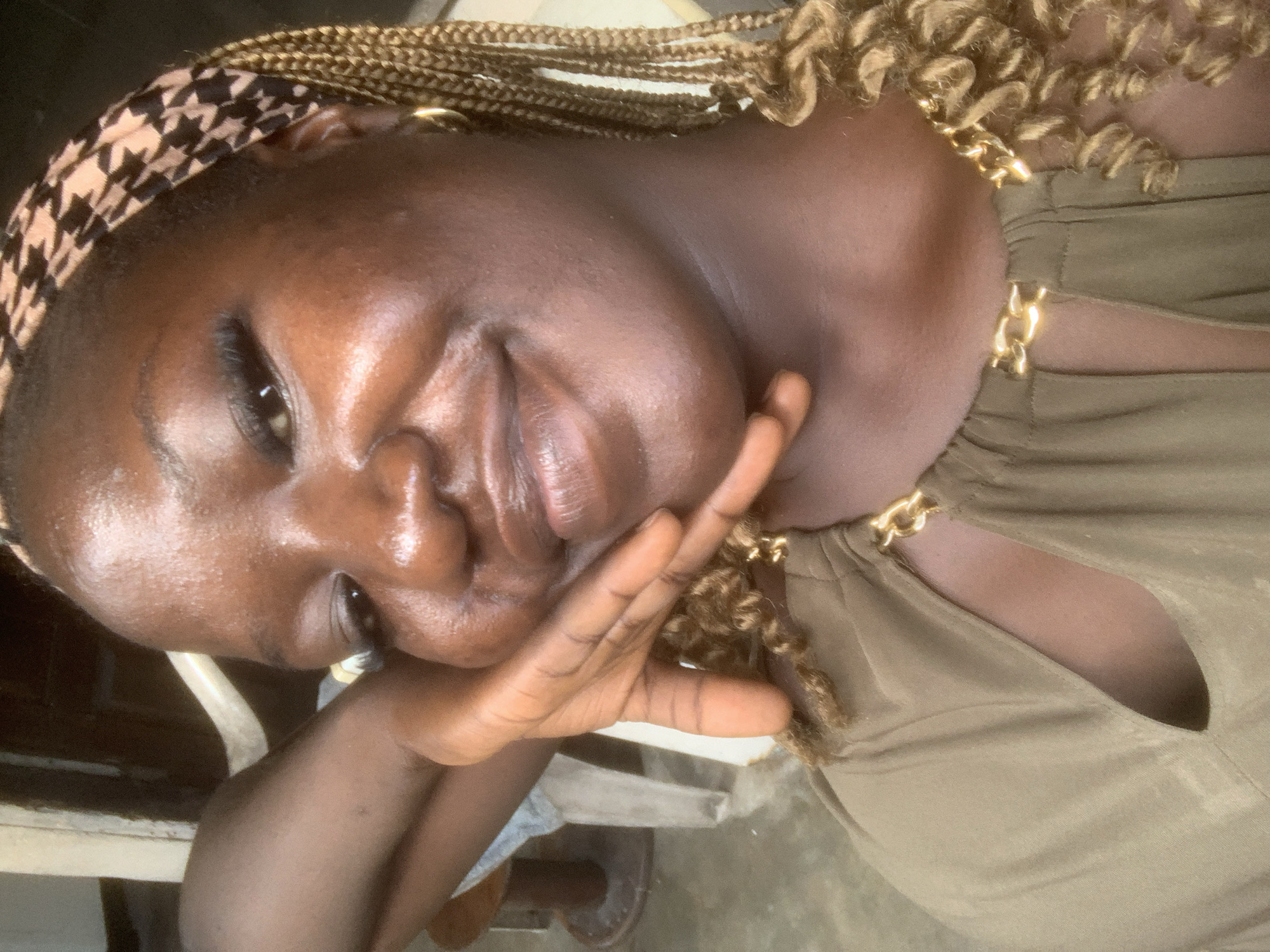 In Eliozu, Nigeria, a close-up, waist-up selfie captures a woman with dark skin and braided hair. She is wearing a khaki-colored garment with a gold chain detail at the neckline. Her left arm is bent, and her hand rests gently on her cheek, with her fingers slightly splayed. She is looking towards the camera with a subtle smile. The background is out of focus but appears to be an indoor setting with muted colors, possibly a wall and furniture. The lighting is soft, highlighting the sheen on her skin.