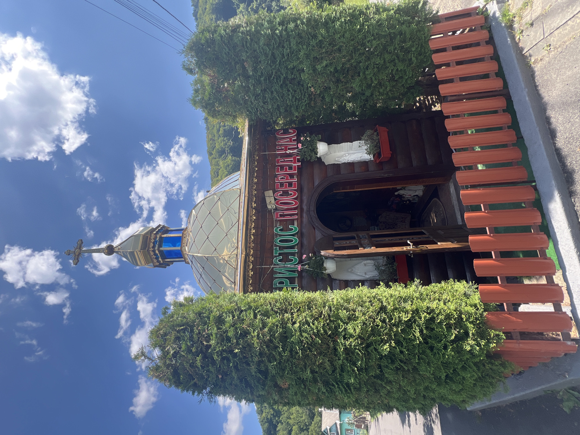 The image captures a small, wooden Orthodox church, likely located in Yaremche, Ukraine, given its architectural style and the surrounding natural landscape. The church features a golden dome topped with a cross, and a blue stained-glass window beneath it. A prominent neon sign in green letters reads "ХРИСТОС ПОСЕРЕДНАС" (Christ is in our midst) across the front facade.
Flanking the entrance are two white statues, one holding a flower. Small potted plants with pink flowers are placed above the statues. The entrance itself is an arched wooden doorway, slightly ajar, revealing steps leading inside. The church is surrounded by a short, orange picket fence.
Two tall evergreen trees stand on either side of the church. In the background, a green, hilly landscape with trees is visible, and a few buildings can be seen to the left, suggesting a semi-rural or suburban setting. The sky is bright blue with scattered white clouds, indicating a sunny day. Power lines are visible in the upper right corner. The overall scene conveys a sense of religious devotion and local community.