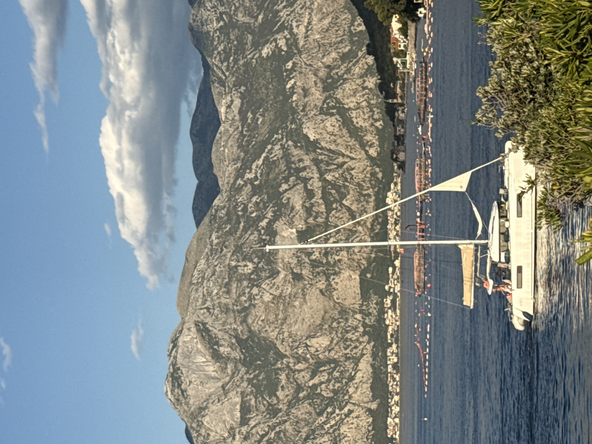 A daytime, eye-level, vertical shot captures a white catamaran sailing on a deep blue body of water. The boat is positioned in the foreground and to the left of the frame, with its sail partially unfurled and a mast extending upwards. A few individuals are visible on the deck of the catamaran. In the background, a town with red-roofed buildings stretches along the coastline. Behind the town, a range of rugged, grey mountains dominates the landscape, their slopes sparsely covered with dark green vegetation. The sky above is a bright blue with scattered white clouds. Several buoys are floating in the water between the town and the catamaran. The image appears to have been taken in the vicinity of Đurići, Montenegro, likely in the Bay of Kotor.